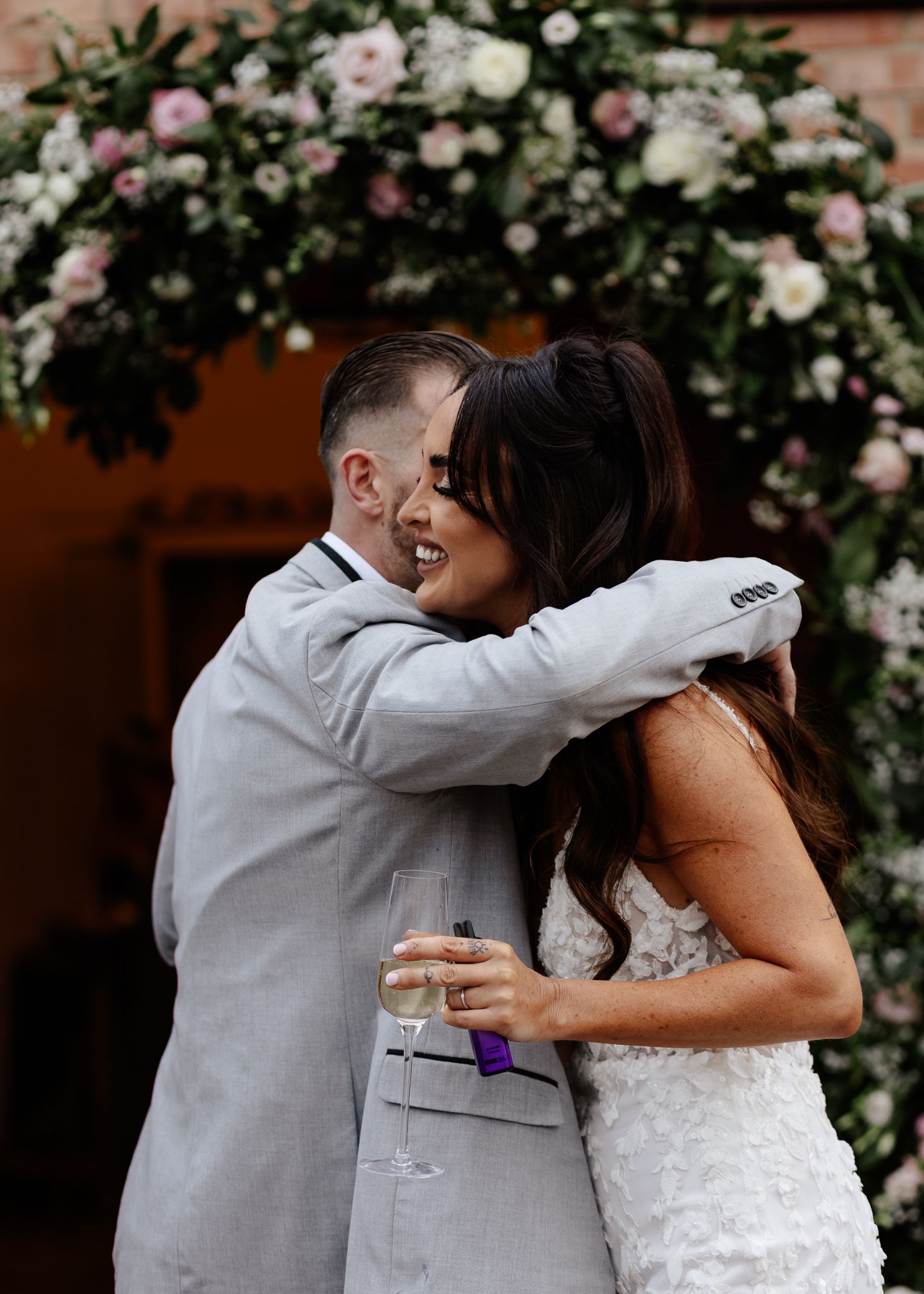 Newlyweds embrace under a floral arch decorated with pink and white roses during their wedding celebration.