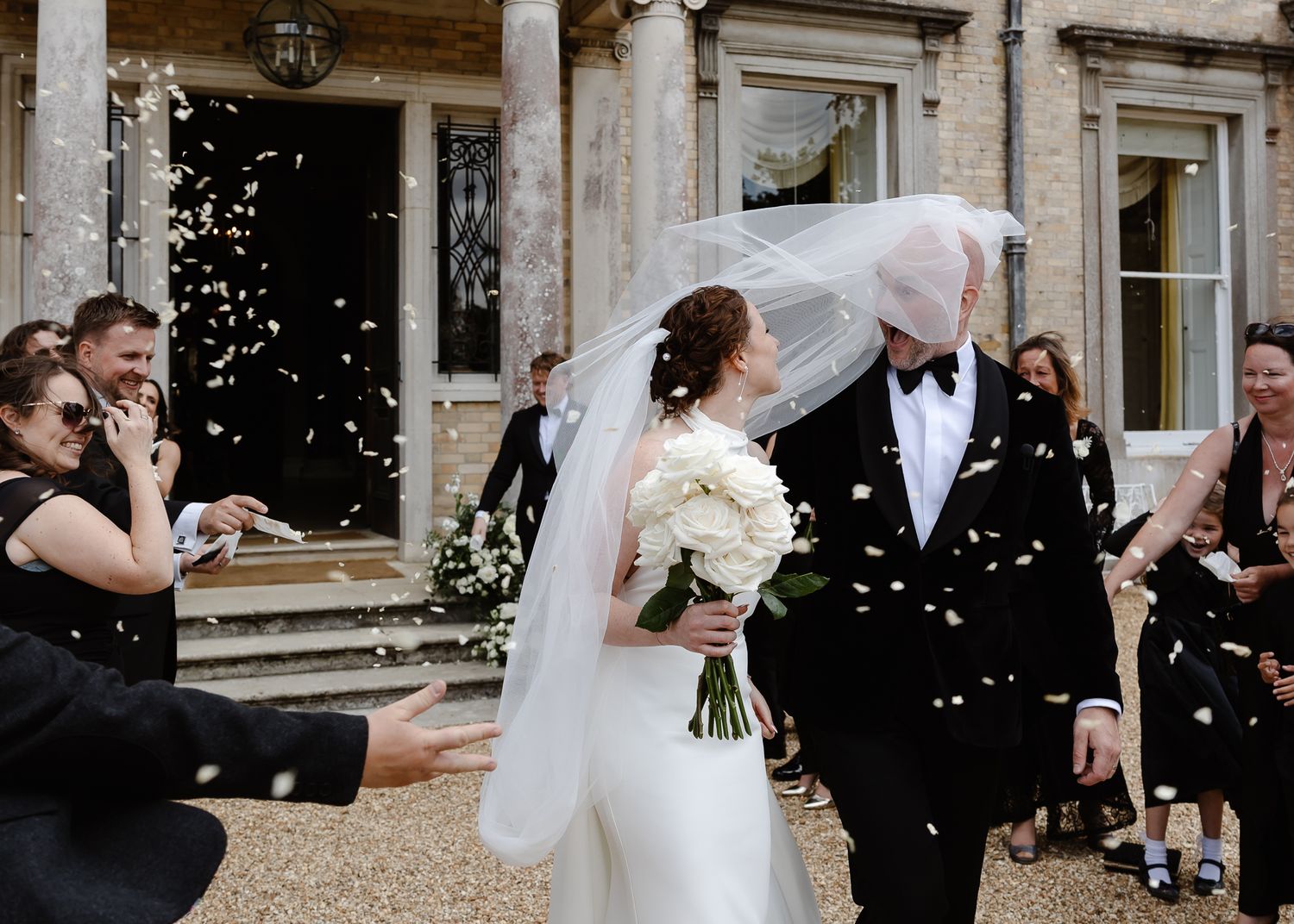 Confetti falls around wedding party members outside a grand stone building entrance.