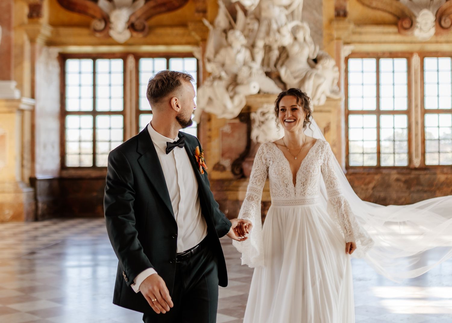 A couple shares an intimate moment in an elegant ballroom with large windows and ornate architectural details.