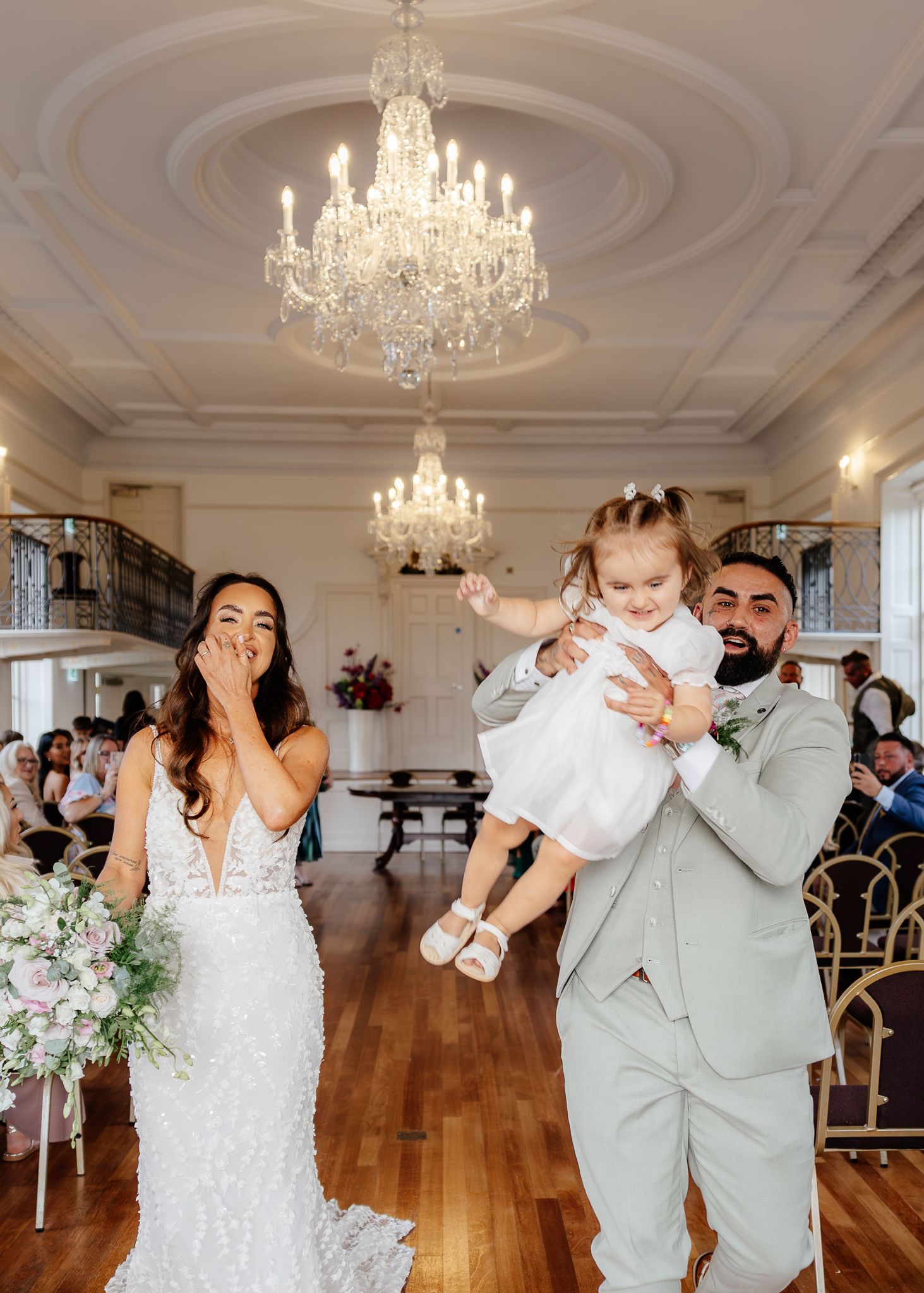 Wedding couple dances joyfully in an elegant ballroom with crystal chandeliers and hardwood floors.