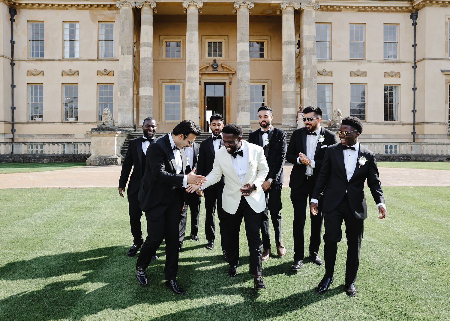 Groomsmen in black tuxedos walk together on a manicured lawn outside an elegant historic building.