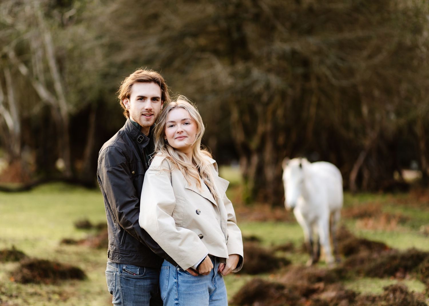 Couple posing outdoors with a white horse in background, surrounded by lush green trees.