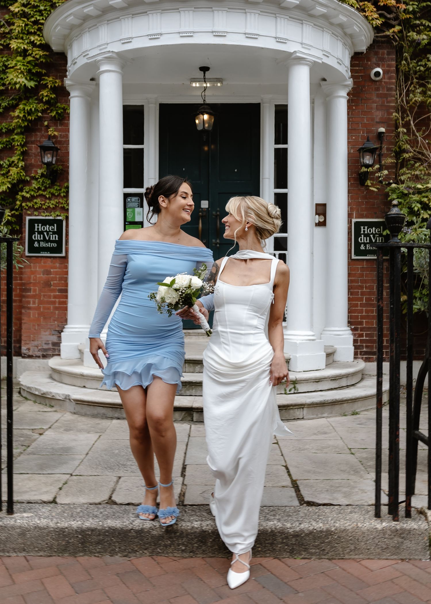Two women in elegant dresses pose outside Hotel du Vin, one in white bridal gown holding bouquet, other in blue dress.