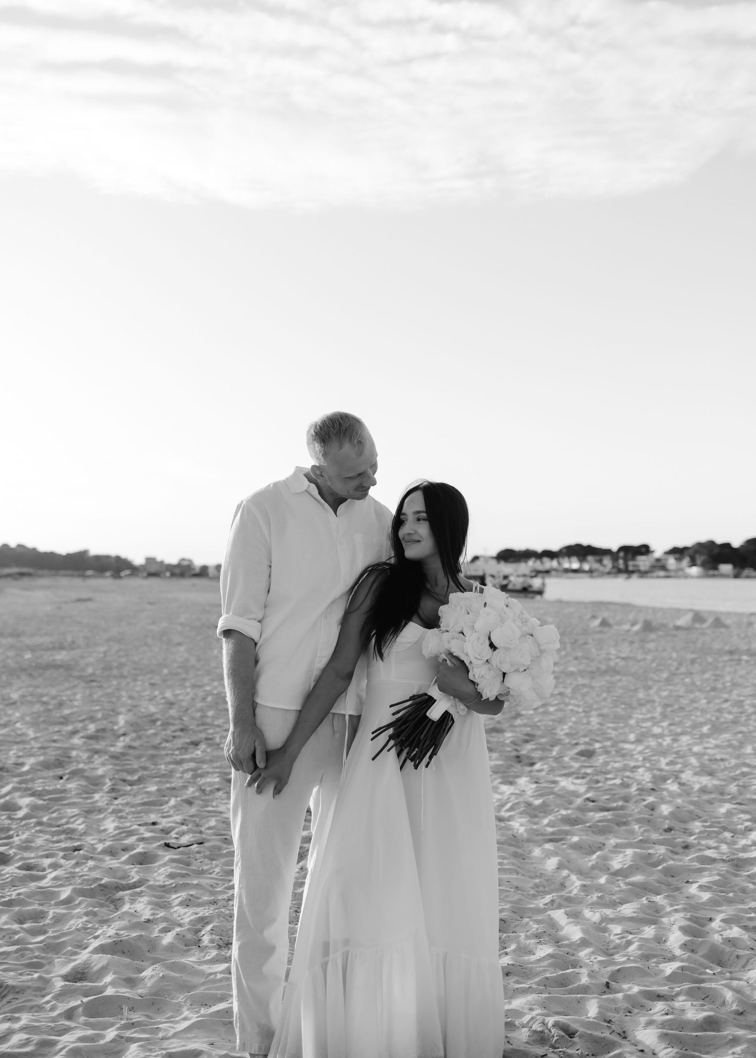 Black and white beach wedding photo of couple, bride in flowing white dress holding bouquet, groom in linen shirt.
