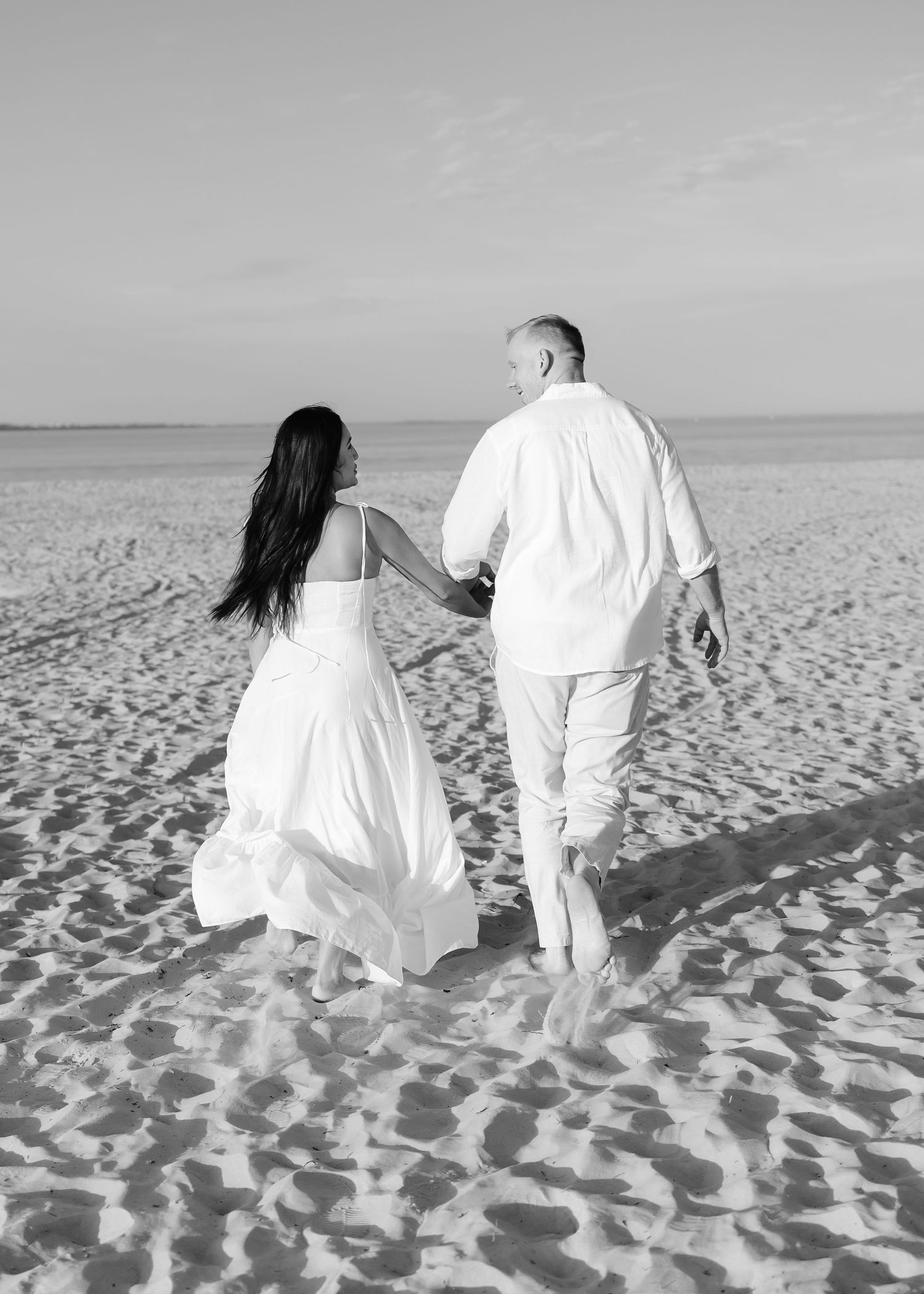 Black and white photo of a couple in white wedding attire holding hands walking on a sandy beach.