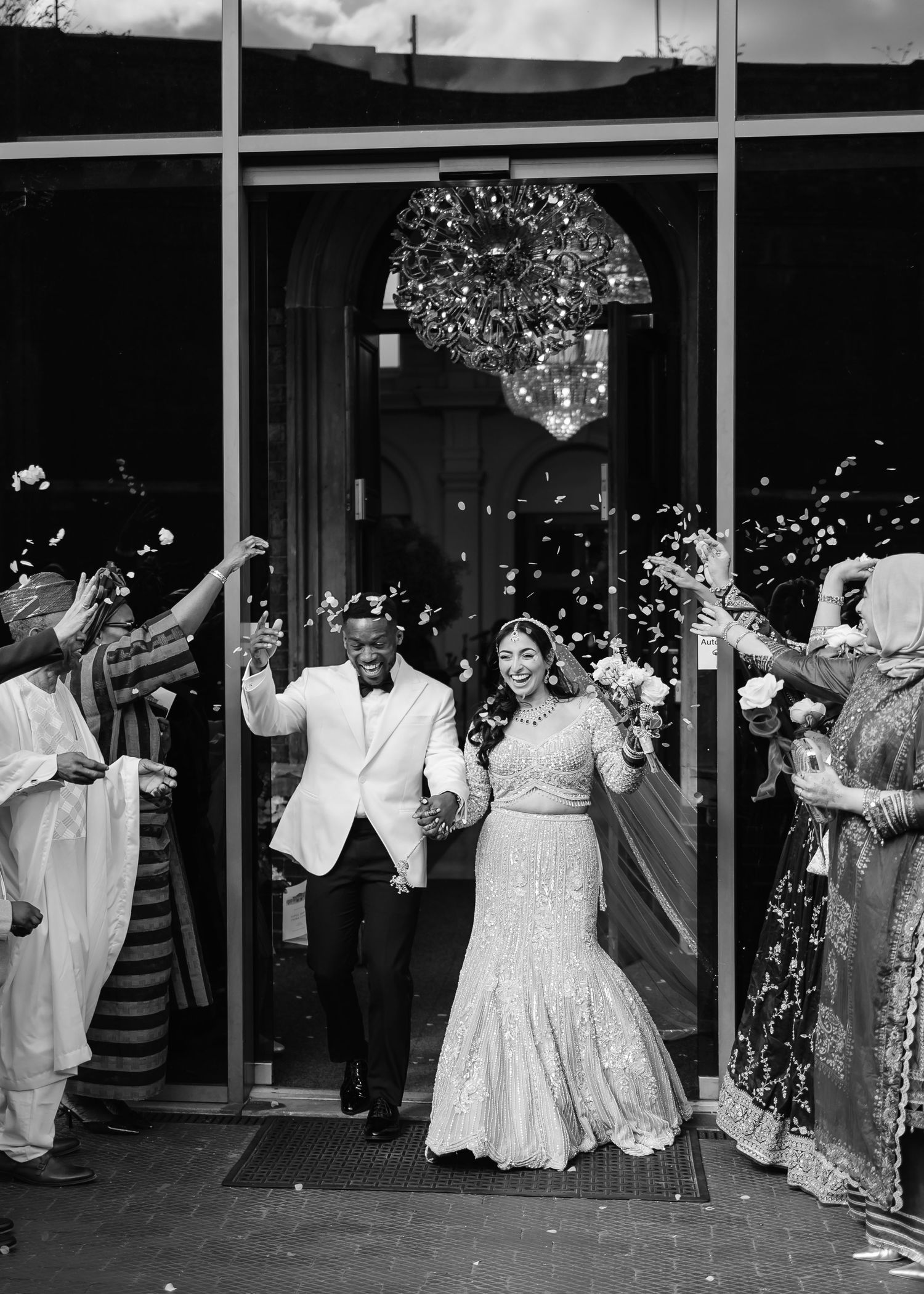 Black and white photo of a joyful couple exiting their wedding venue as guests toss confetti in celebration.