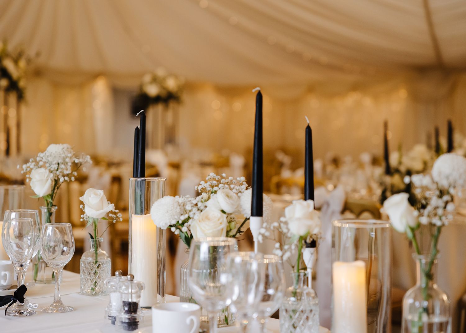Elegant wedding reception table settings with white floral centerpieces, glowing candles and crystal glassware in a tent.
