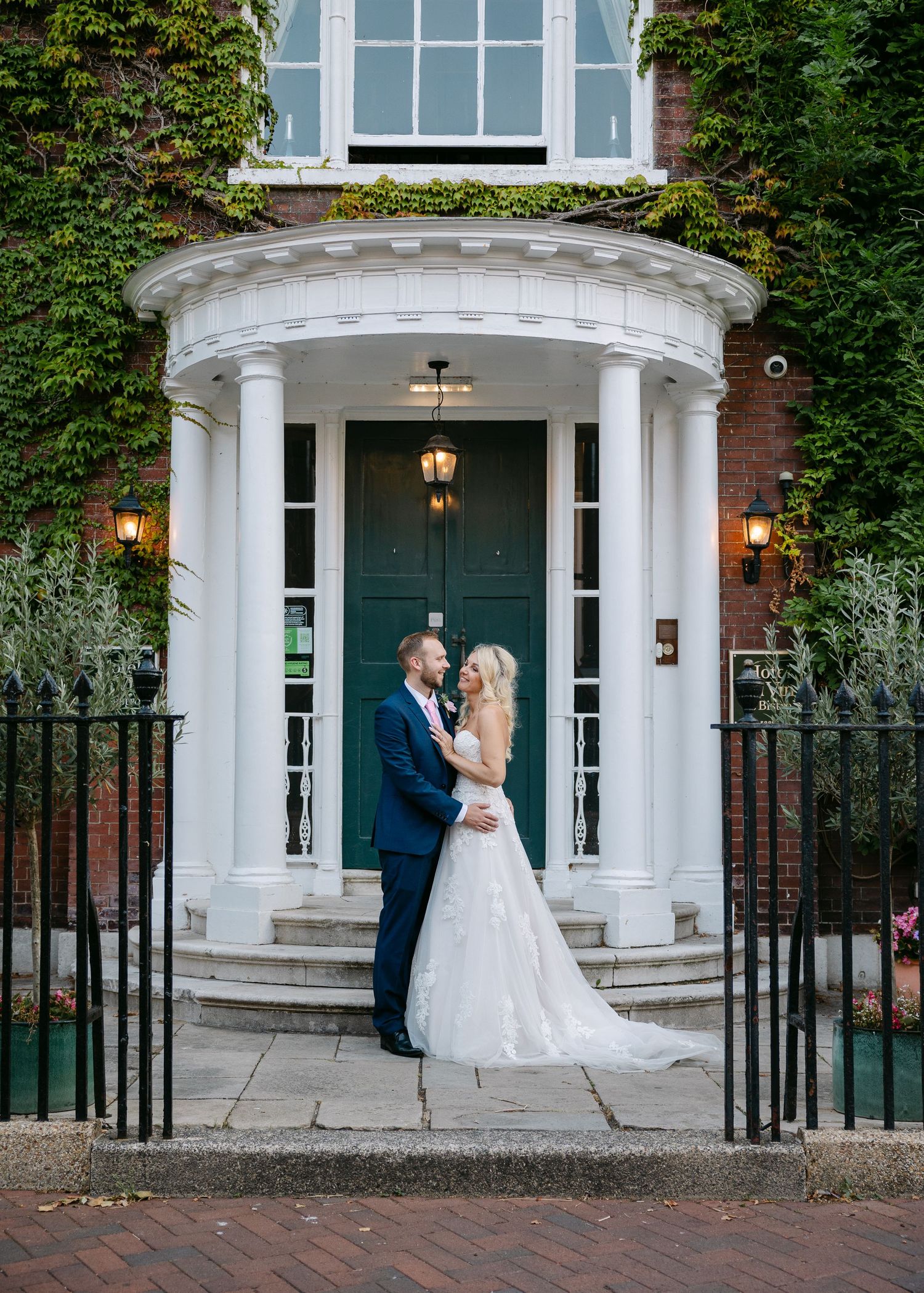 Bride and groom embrace before ivy-covered brick building with white columns and teal door on their wedding day.