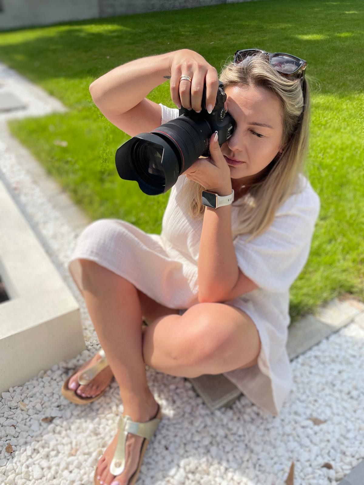 A photographer in a white top sits on concrete steps while taking photos with a professional camera outdoors.