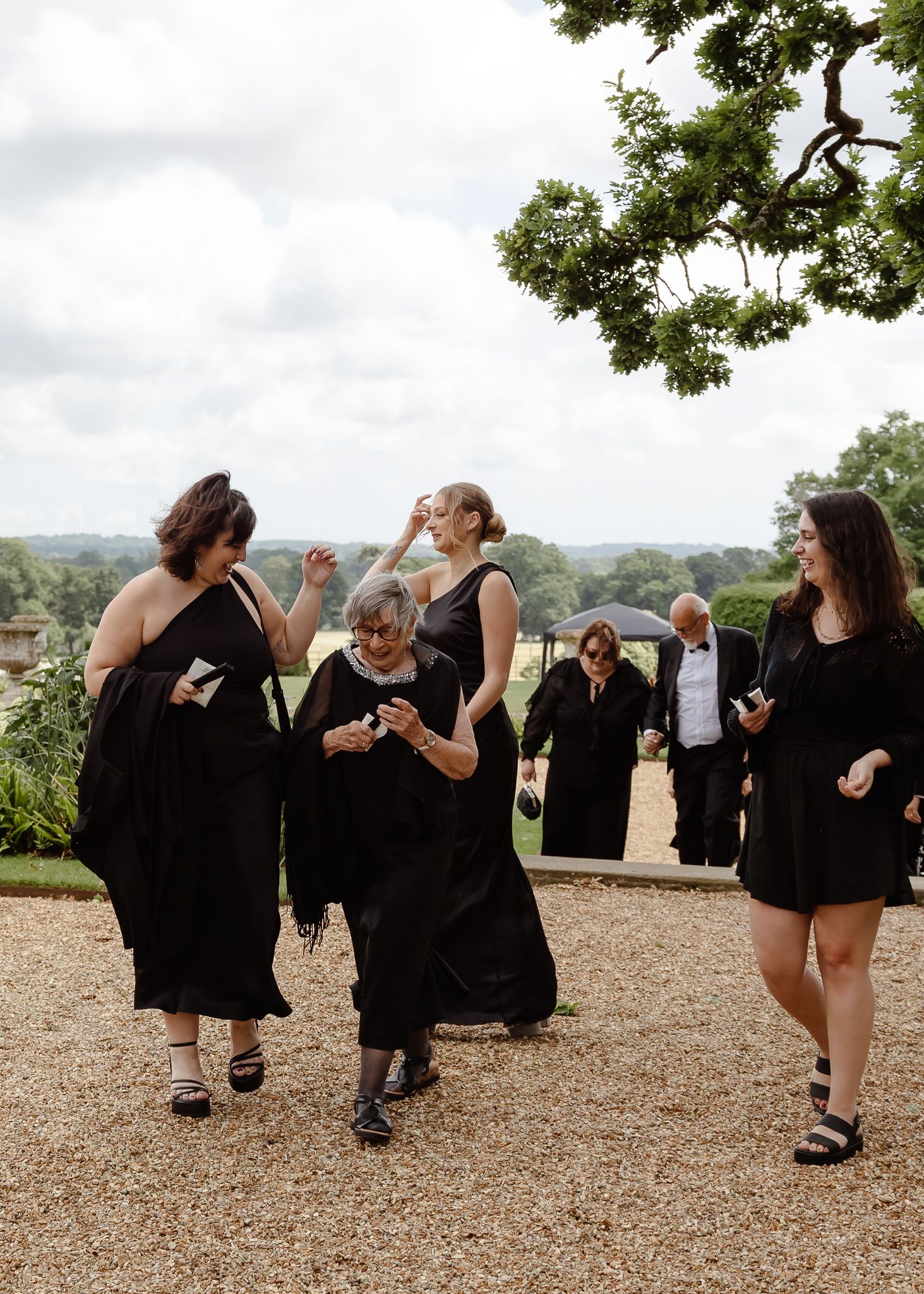 Wedding party in black attire poses playfully on gravel path at elegant venue.