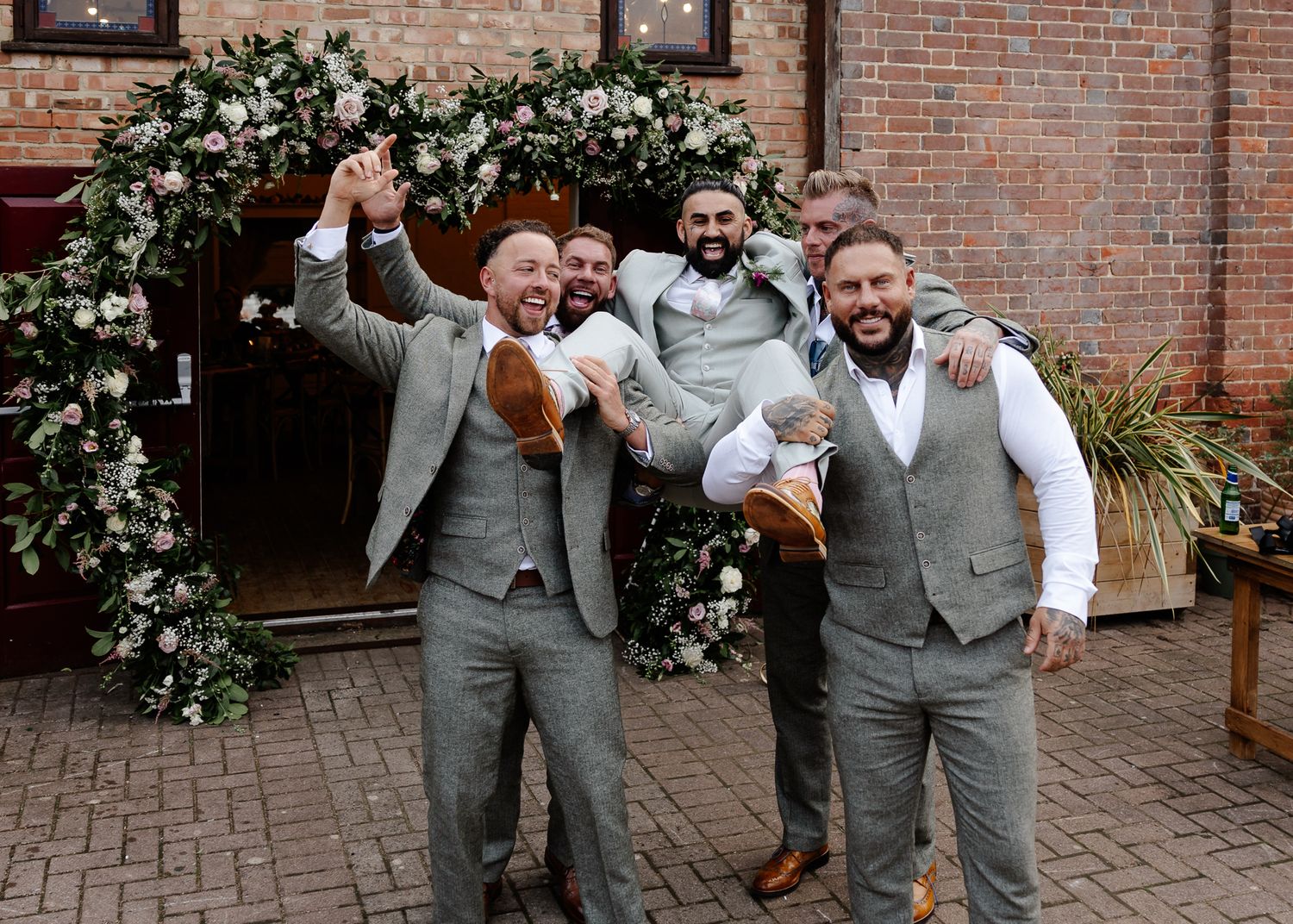A group of groomsmen in light gray vests celebrate joyfully in front of a brick wall adorned with white floral decorations.