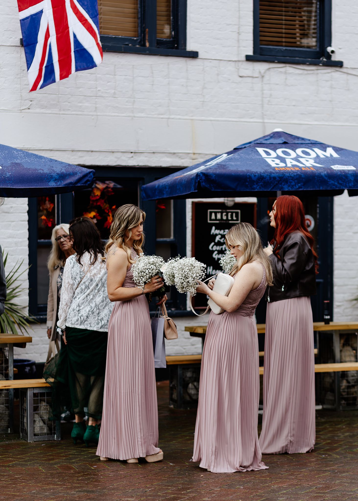 A group in pink dresses and patterned tops stand together at an outdoor venue decorated with British Union Jack flags.