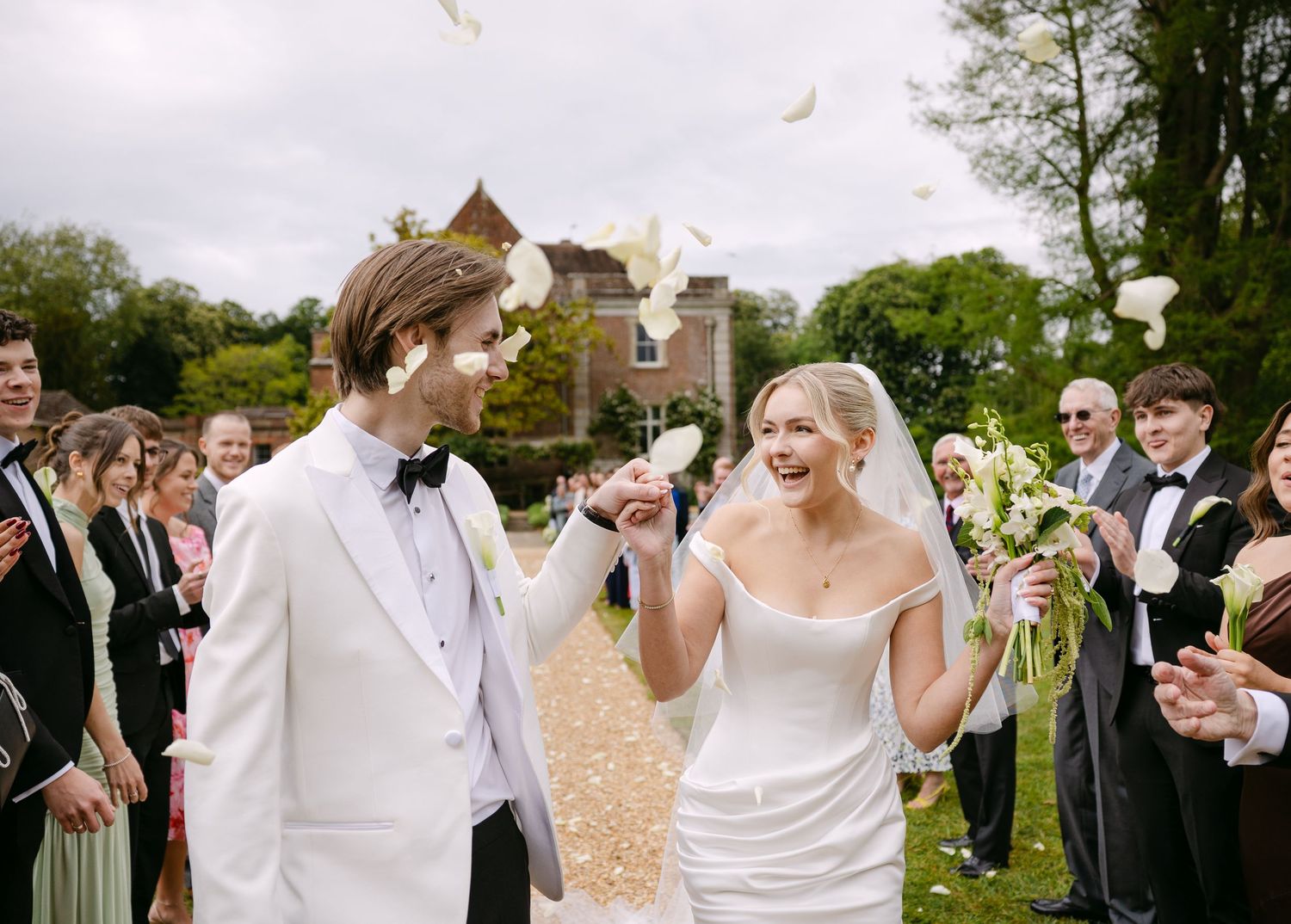 Joyful bride and groom walk through petal confetti shower at outdoor wedding ceremony near historic stone building.