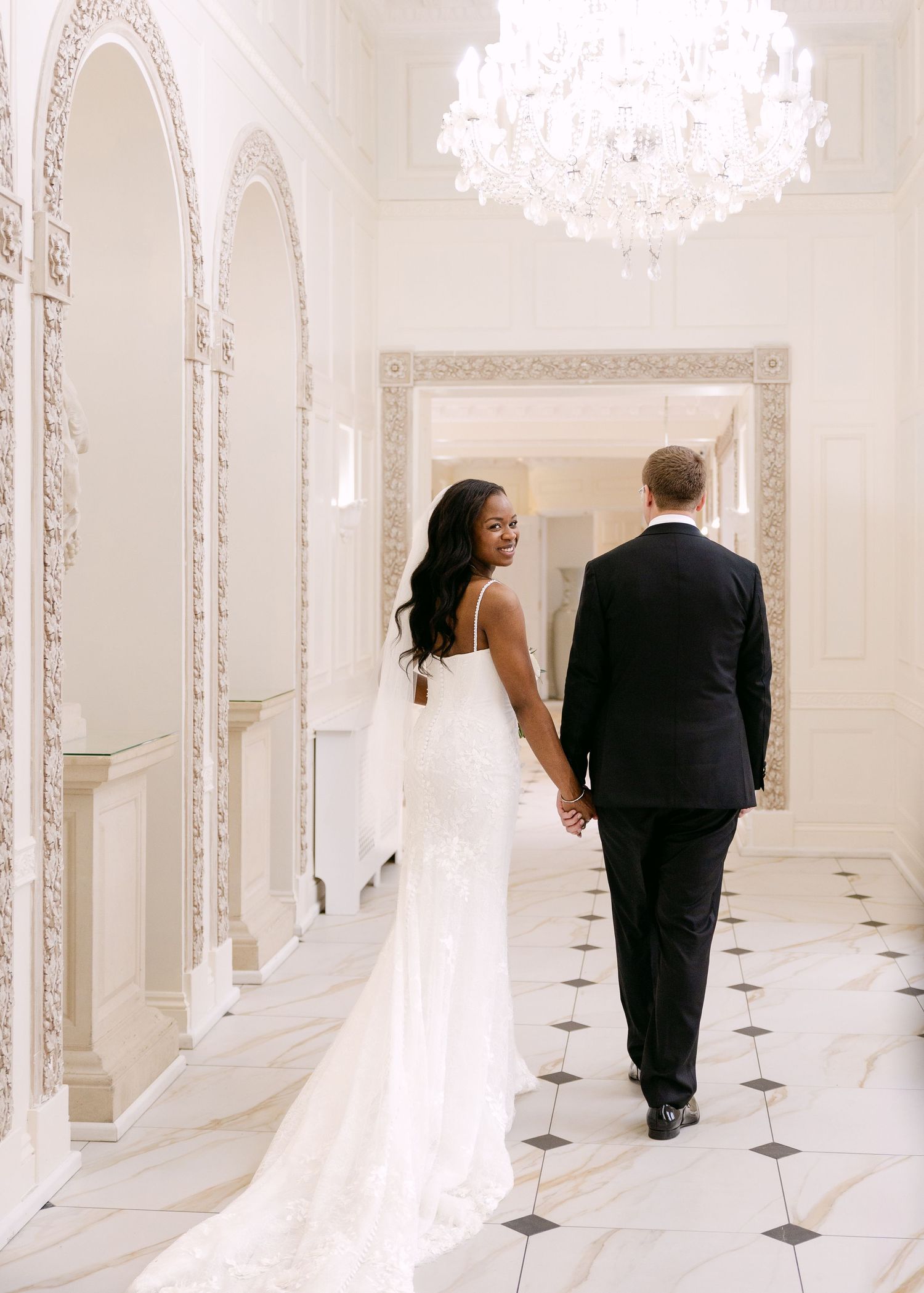 Bride and groom walking hand in hand through elegant white marble hallway with crystal chandelier.