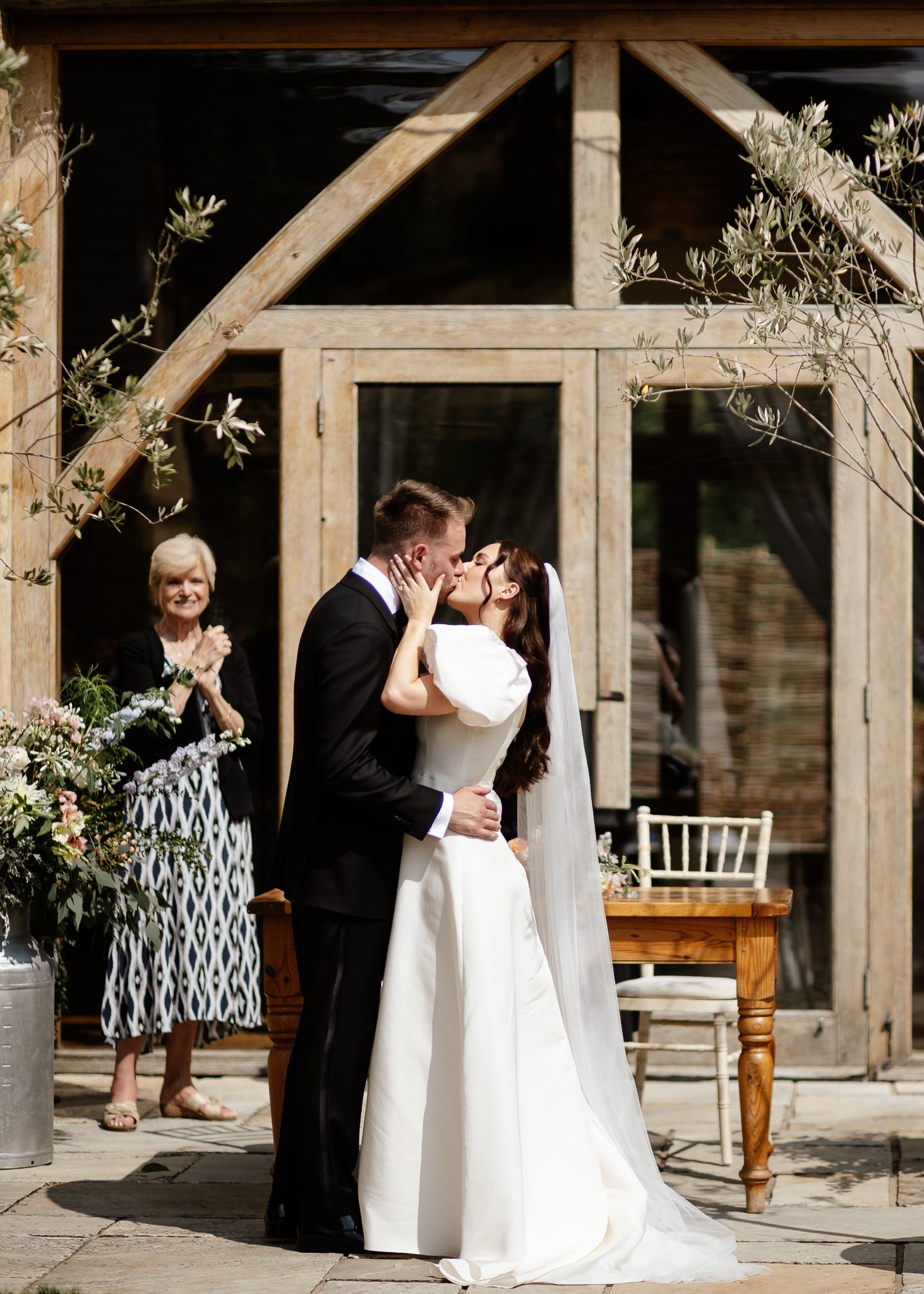 Wedding couple shares a romantic kiss in an elegant outdoor courtyard with wooden beams and white chairs.