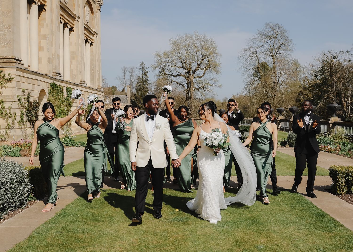 Wedding party members pose playfully in a line on a green lawn outside an elegant stone building.