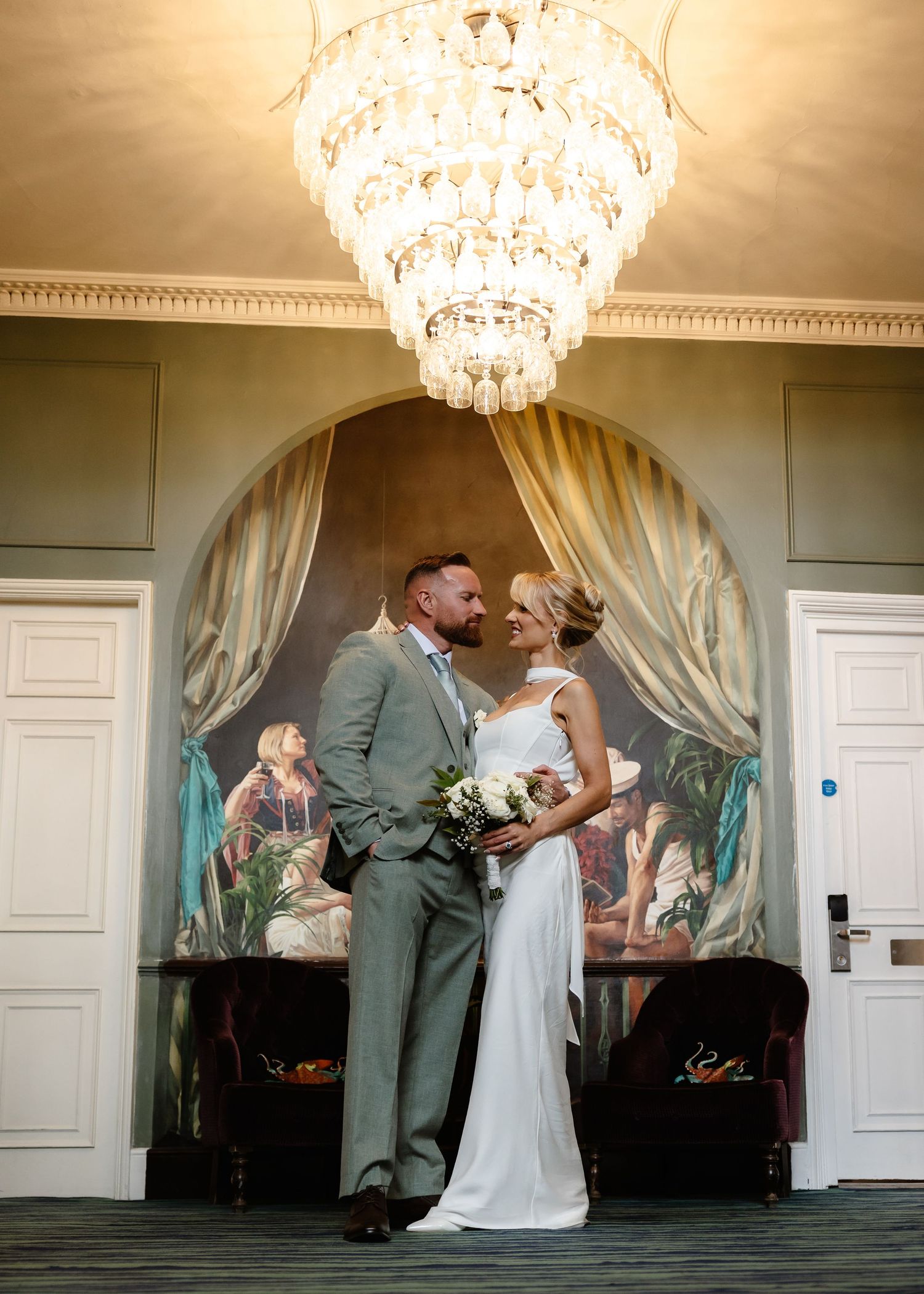 Bride and groom share a romantic moment under a crystal chandelier beside an elegant arched mural.