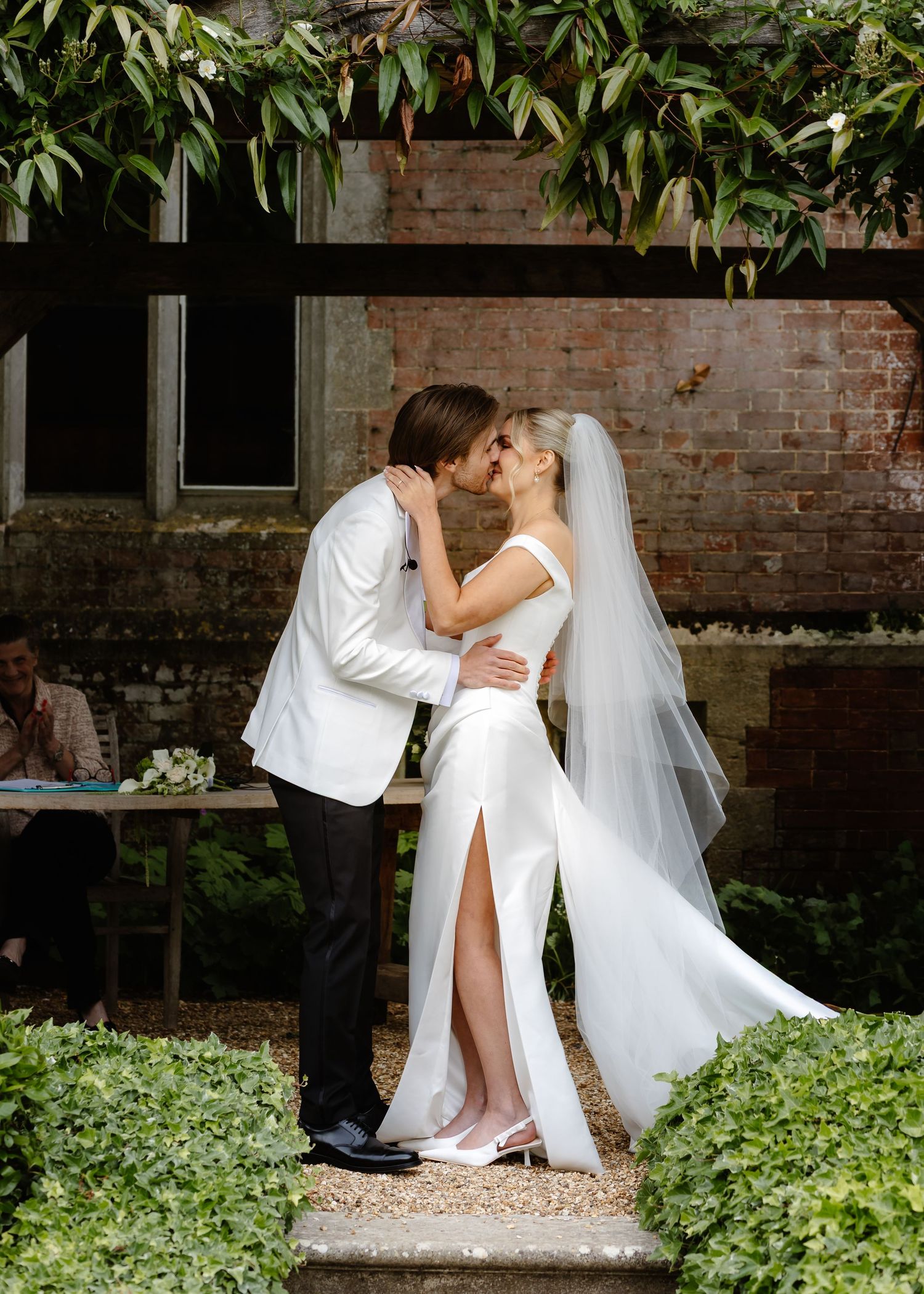A bride and groom share their first kiss at an outdoor wedding ceremony surrounded by lush greenery and a rustic brick wall.