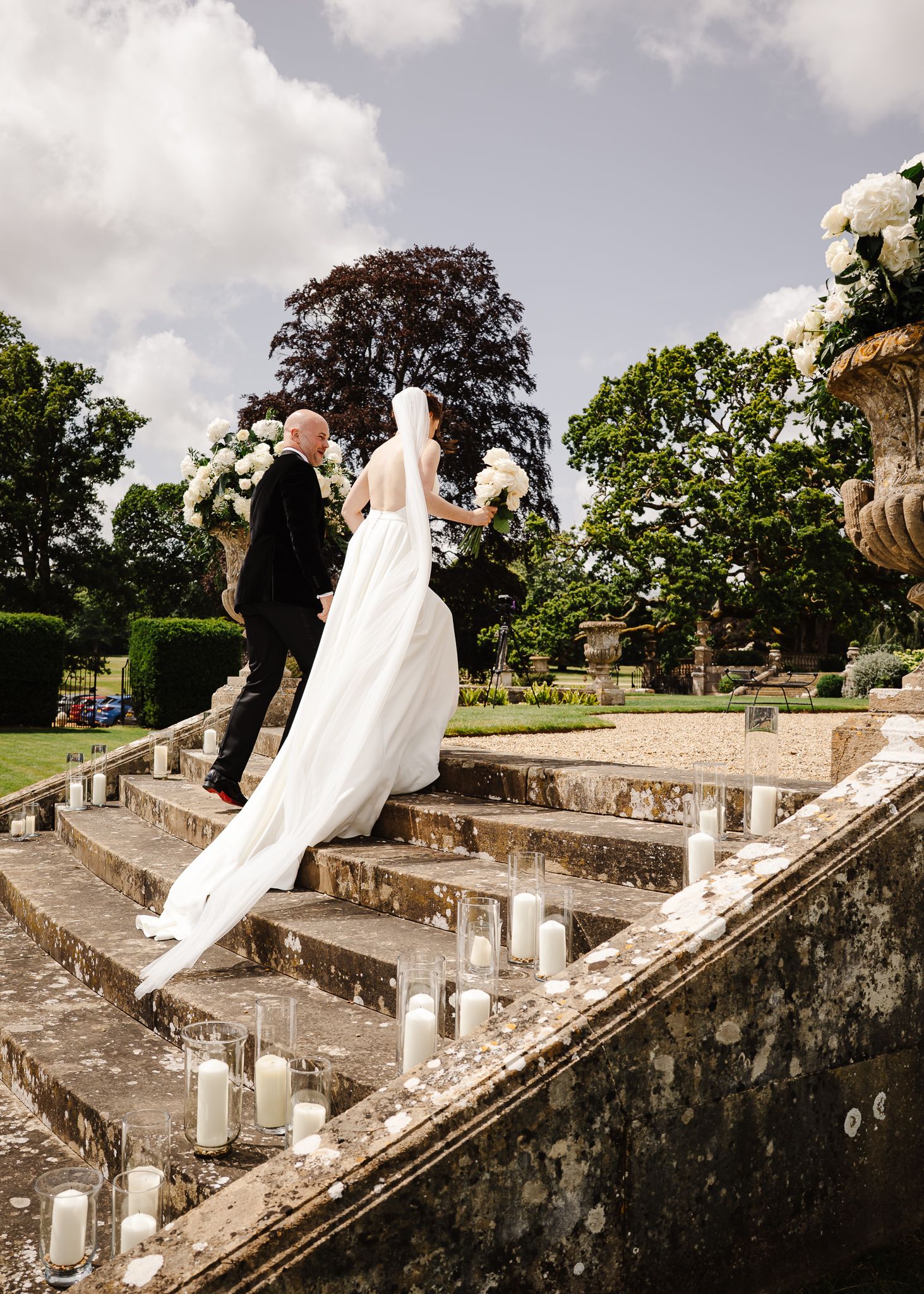 A couple ascends elegant stone steps decorated with candles at an outdoor garden wedding ceremony.