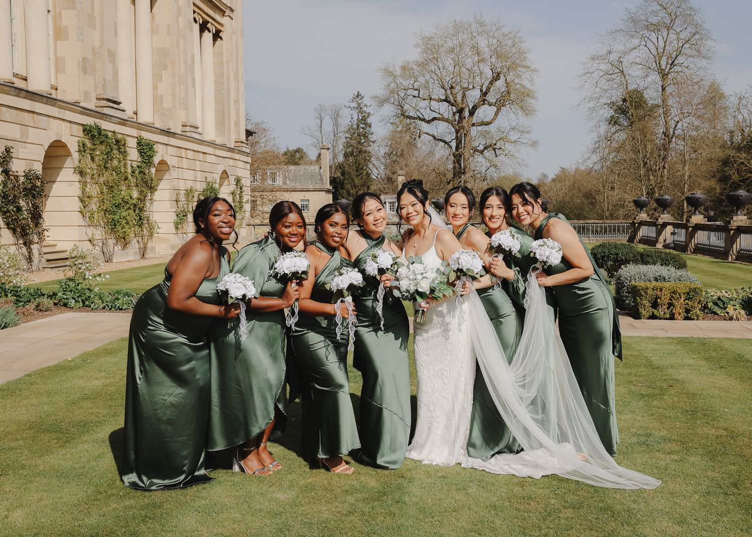 Wedding party in green dresses and formal attire poses together on grand estate grounds.