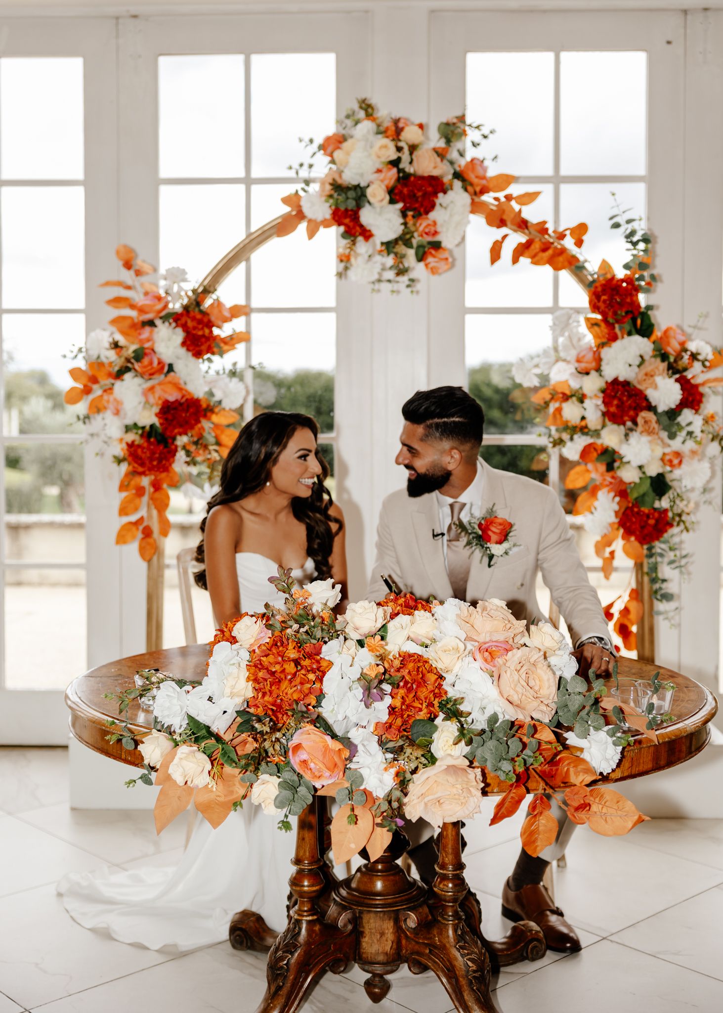 Orange and white floral arrangements with autumn colors displayed in a bright white room with large windows.
