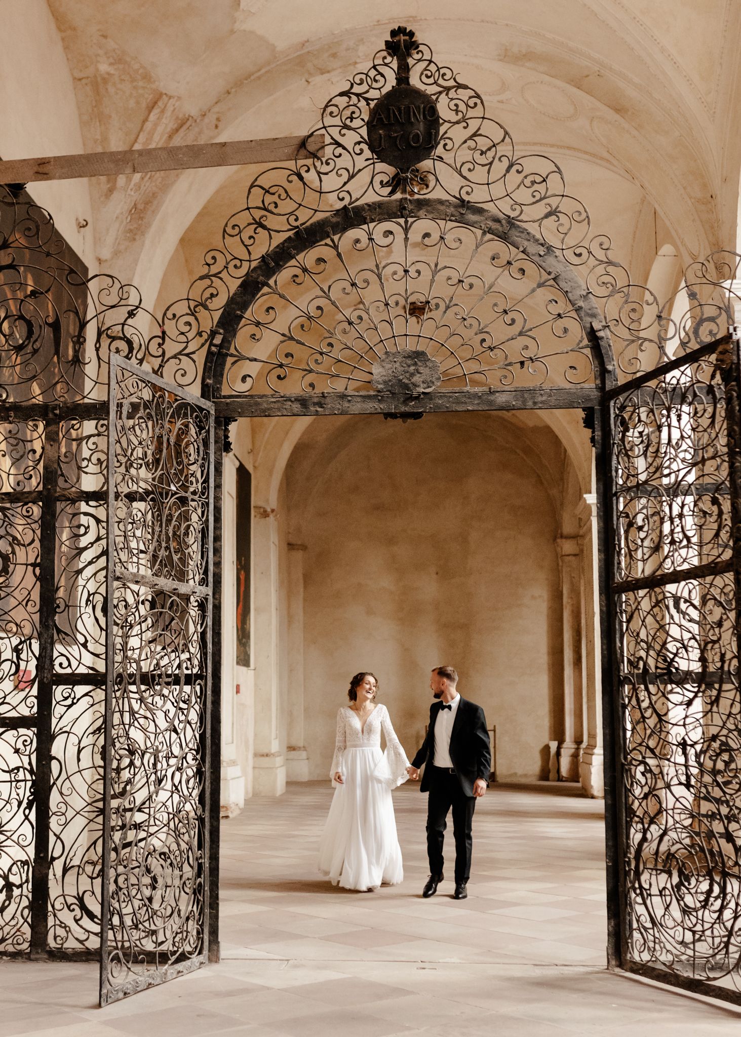 A couple stands beneath an elaborate wrought iron archway in a classic European architectural setting.