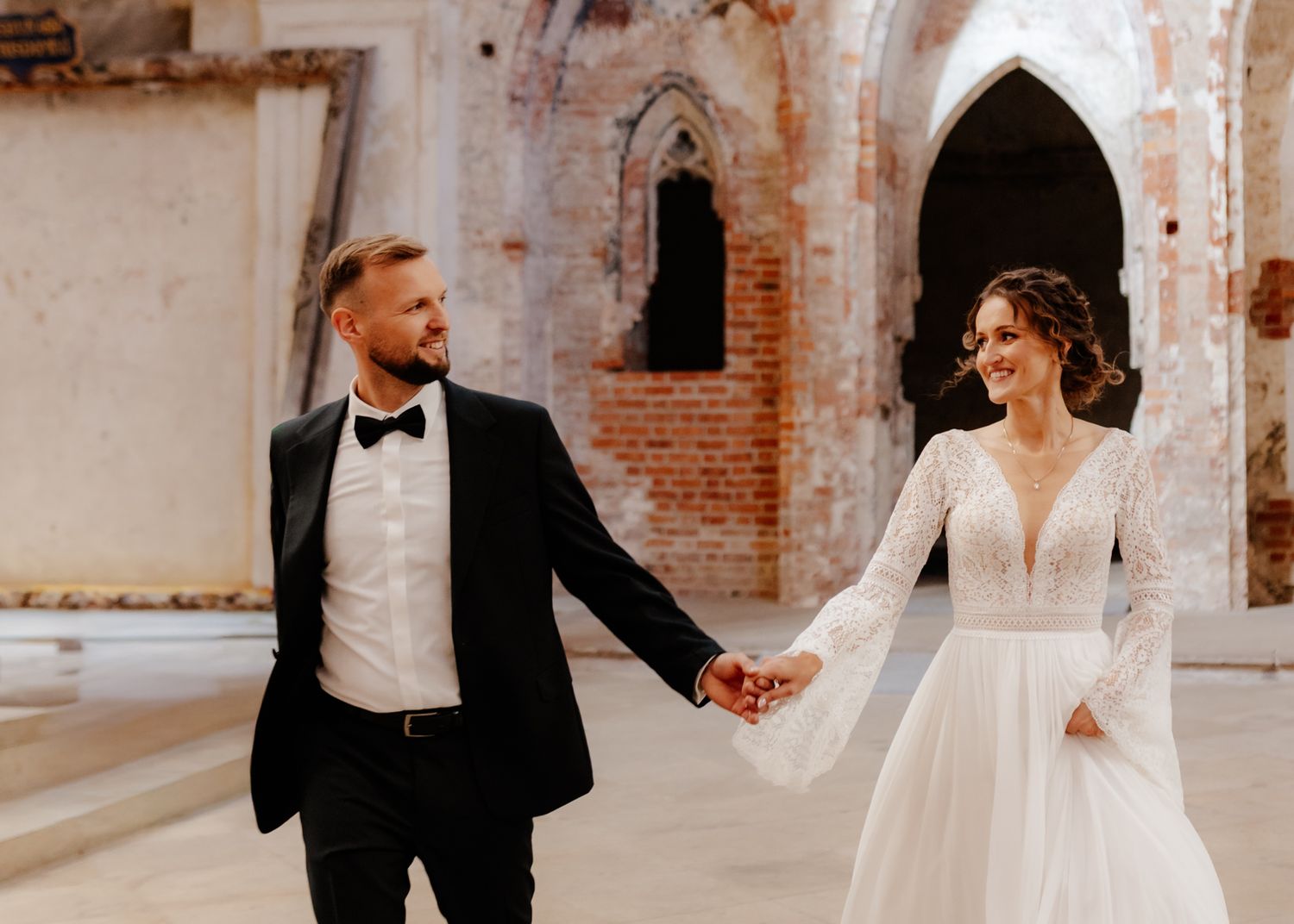 A joyful wedding couple walks hand-in-hand through a rustic brick archway in warm sunlight.
