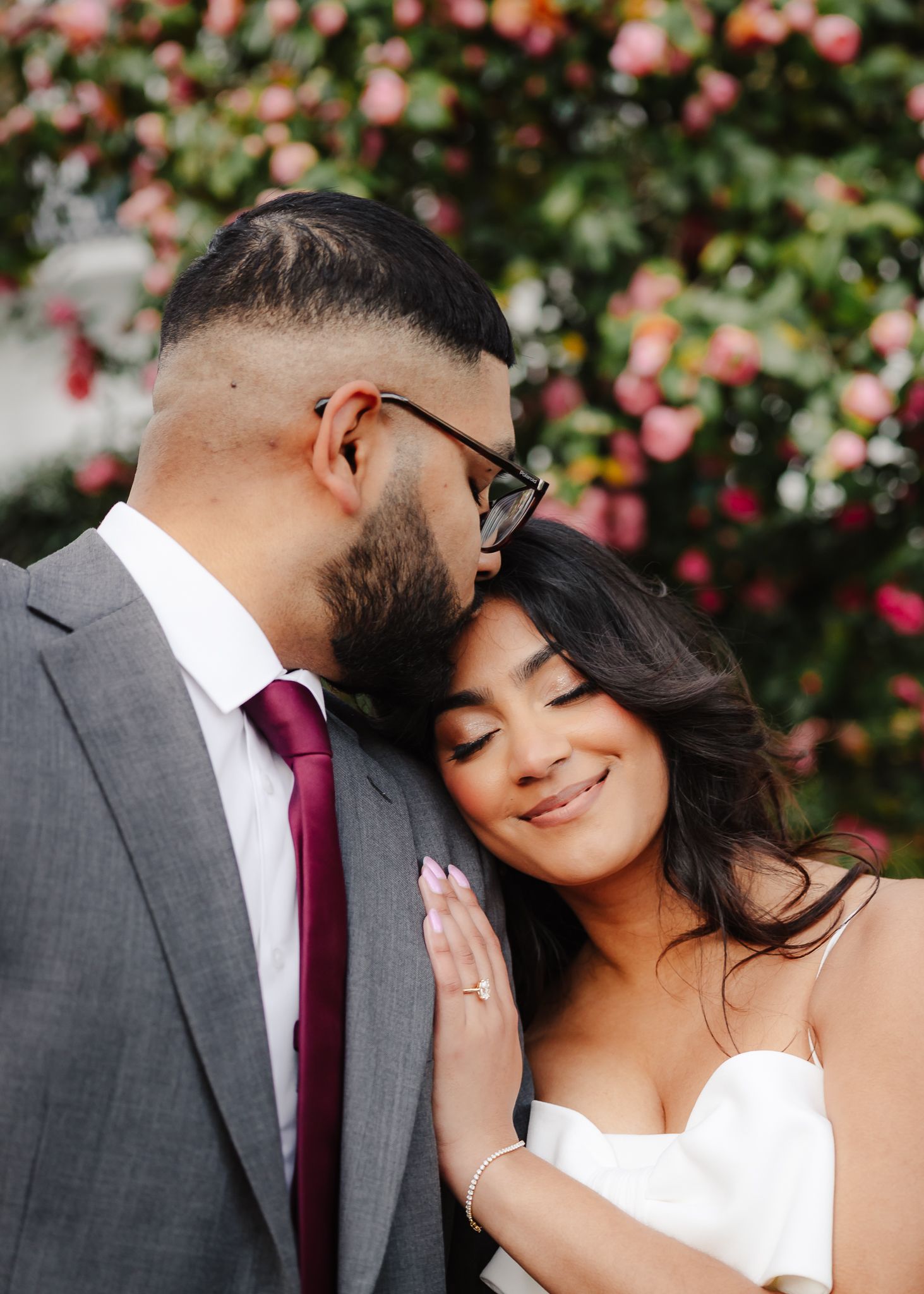 A romantic wedding couple embraces in front of pink flowers, with the bride in white and groom in grey suit and red tie.