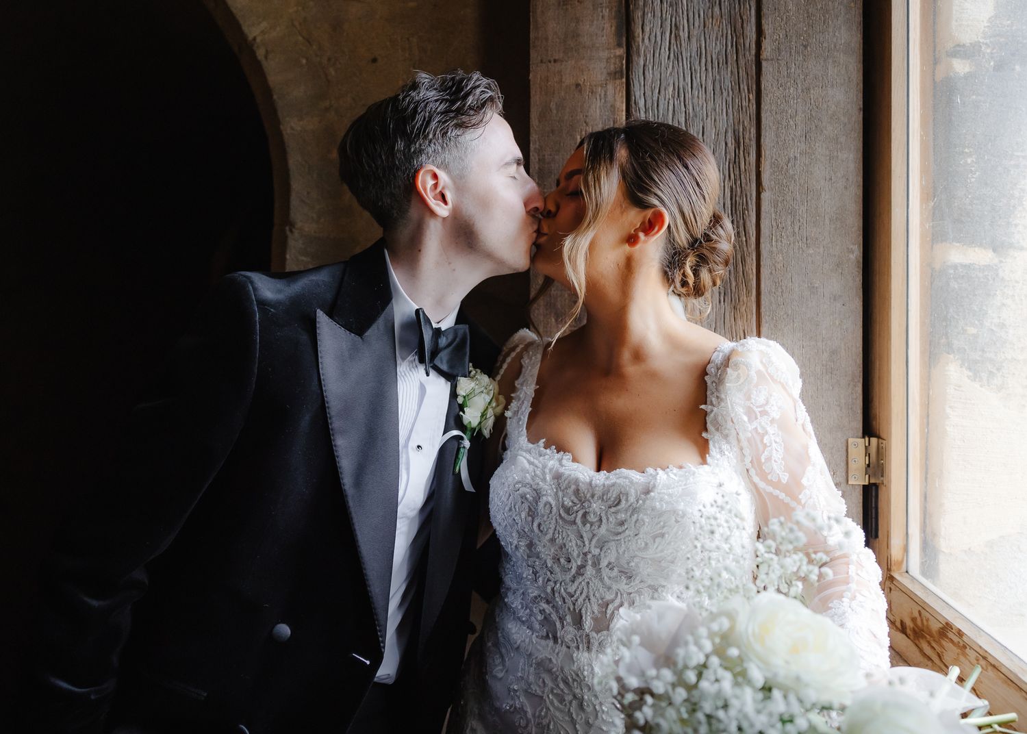 A romantic wedding couple stands near a window in dramatic lighting with the bride wearing a lace gown.