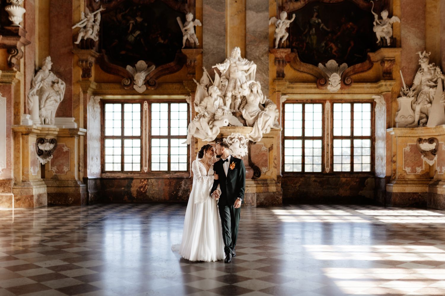 An elegant baroque ballroom with ornate ceiling decorations and large windows letting in natural light.