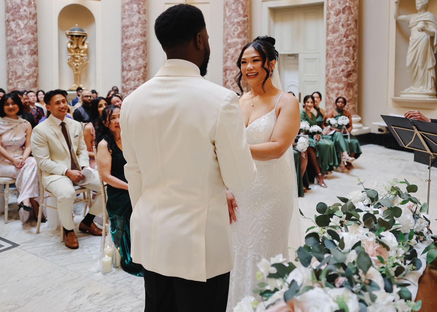 A wedding celebration in an elegant marble courtyard with pink walls and white-clad guests mingling among floral decorations.