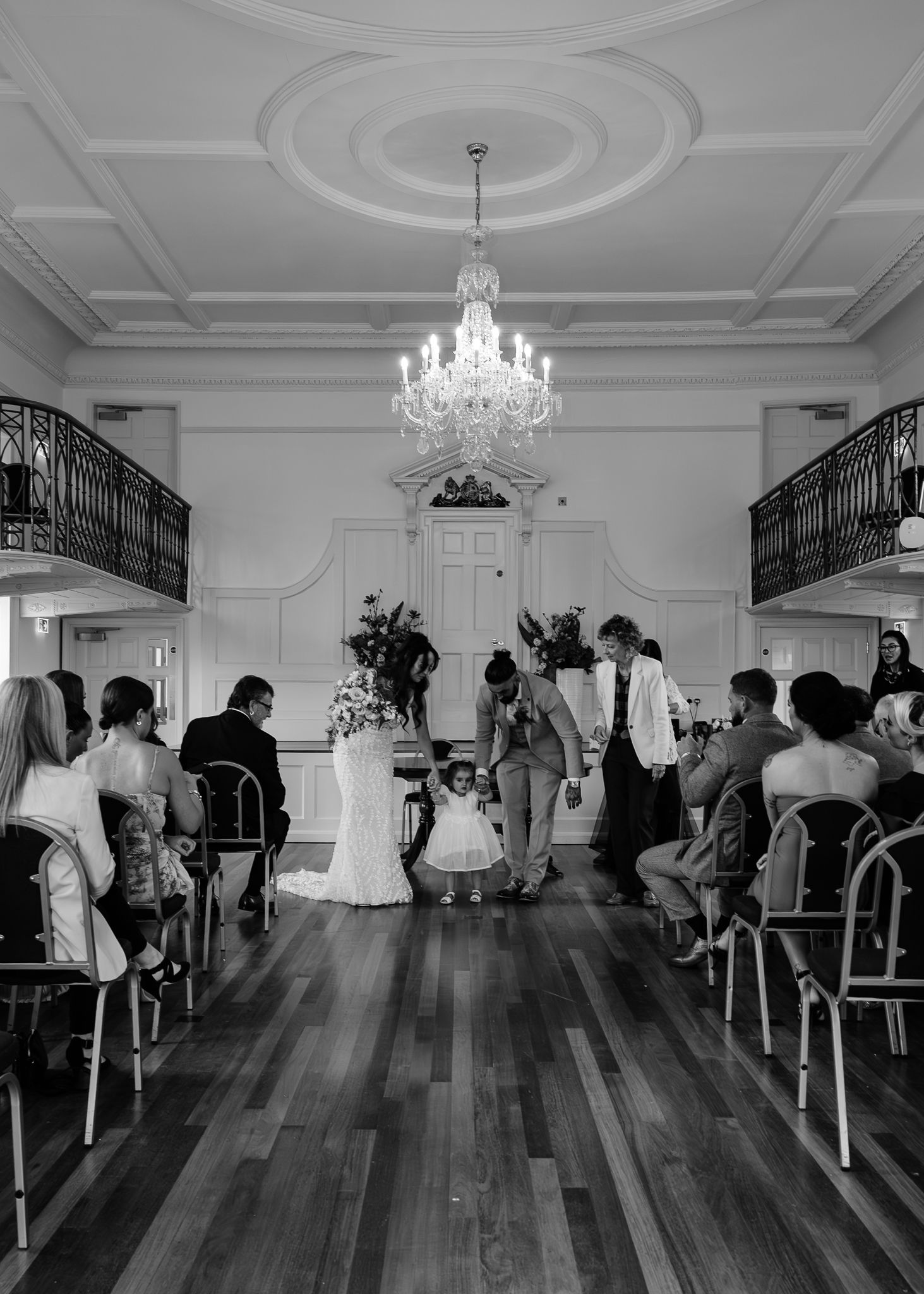 A panoramic view of a wedding ceremony in an elegant white ballroom with crystal chandeliers and hardwood floors.