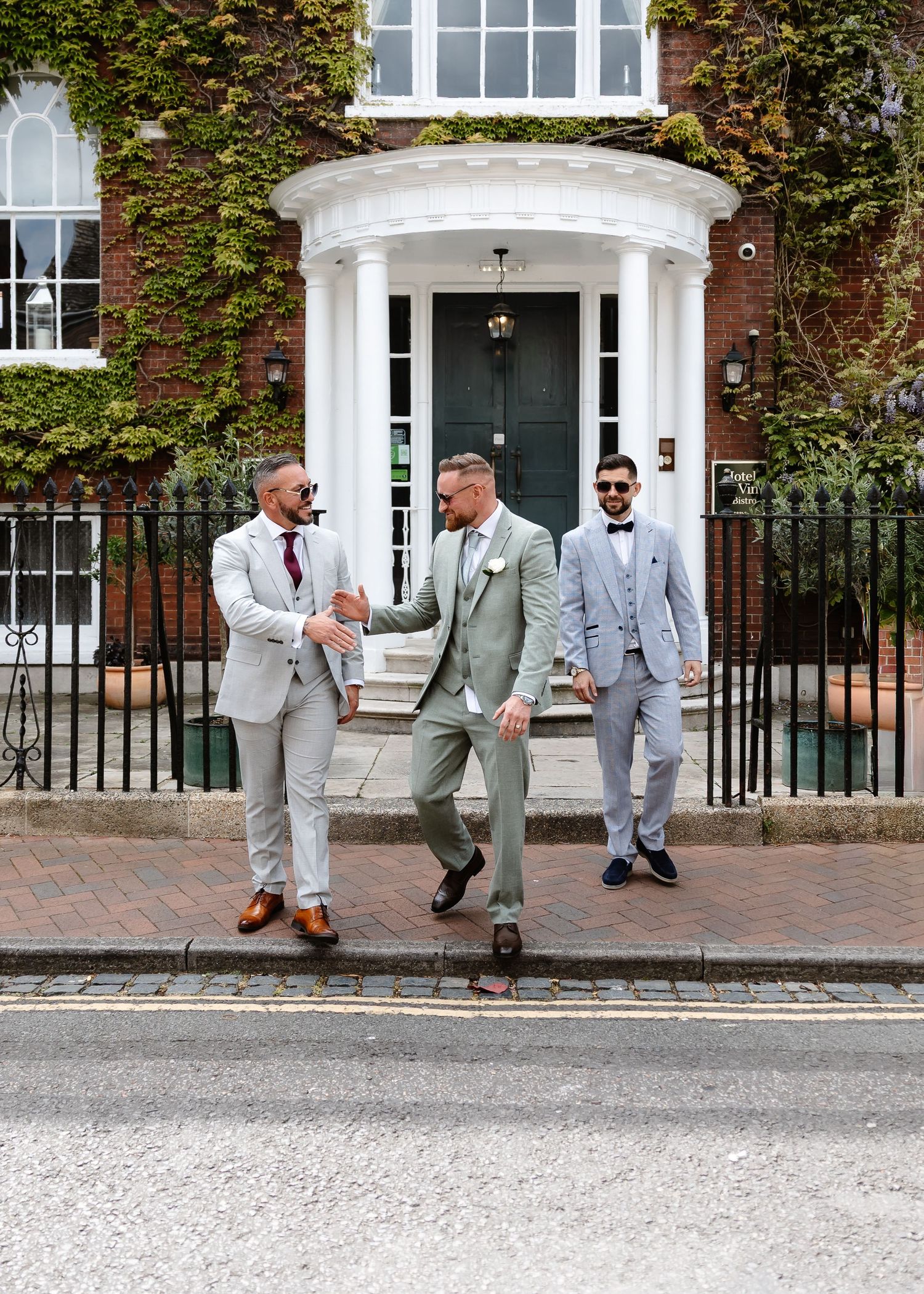 Three groomsmen in sage green and grey suits pose outside an ivy-covered brick building on a wedding day.