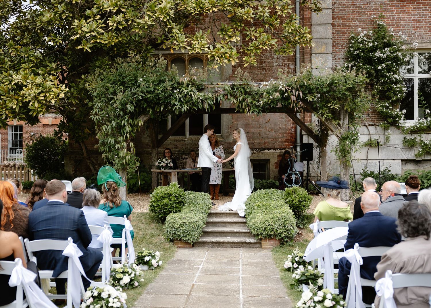 Outdoor wedding ceremony at a brick manor house with guests seated in white chairs along a floral aisle.