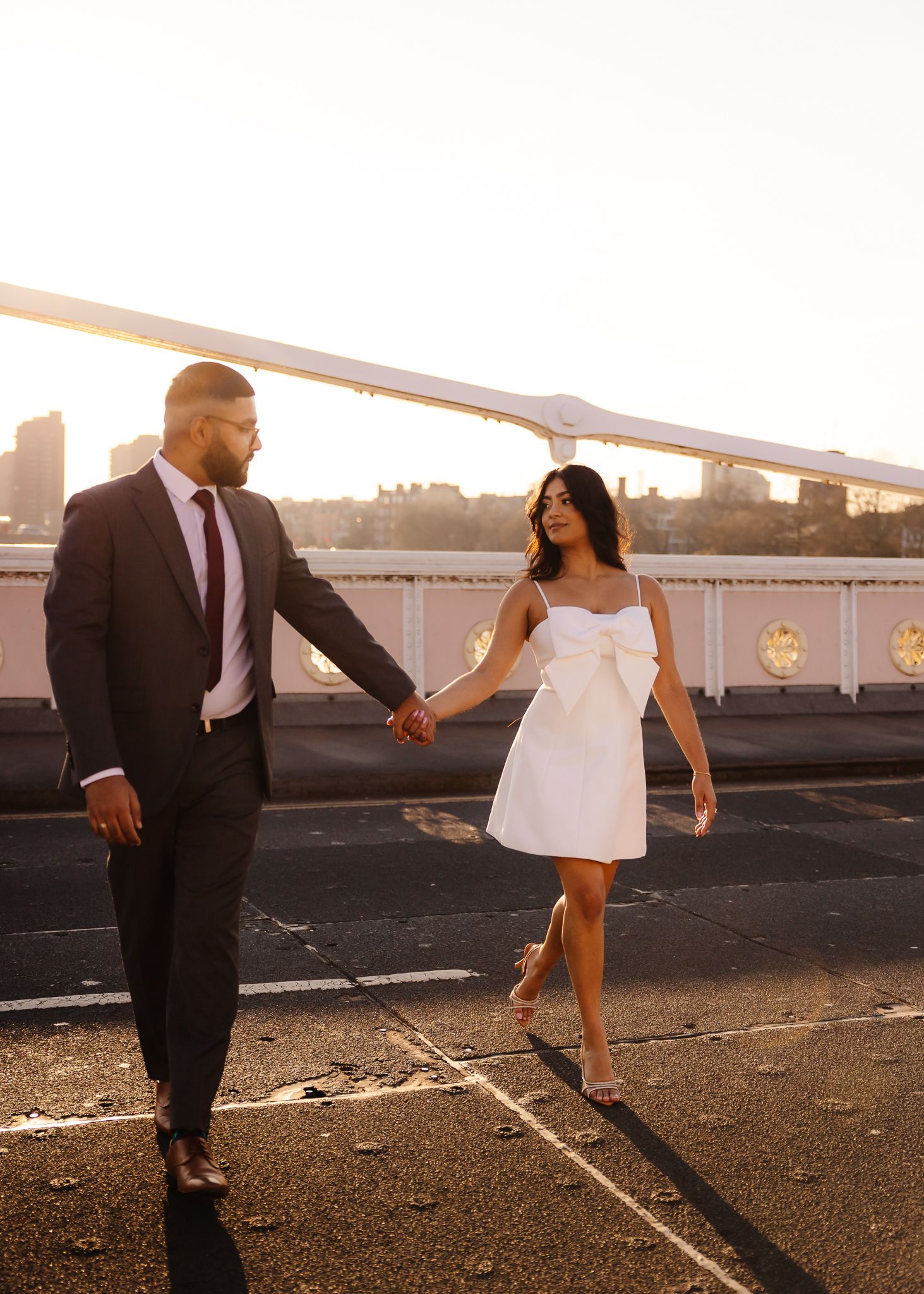 Couple walking hand in hand on a bridge at sunset, man in dark suit and woman in white bow mini dress.
