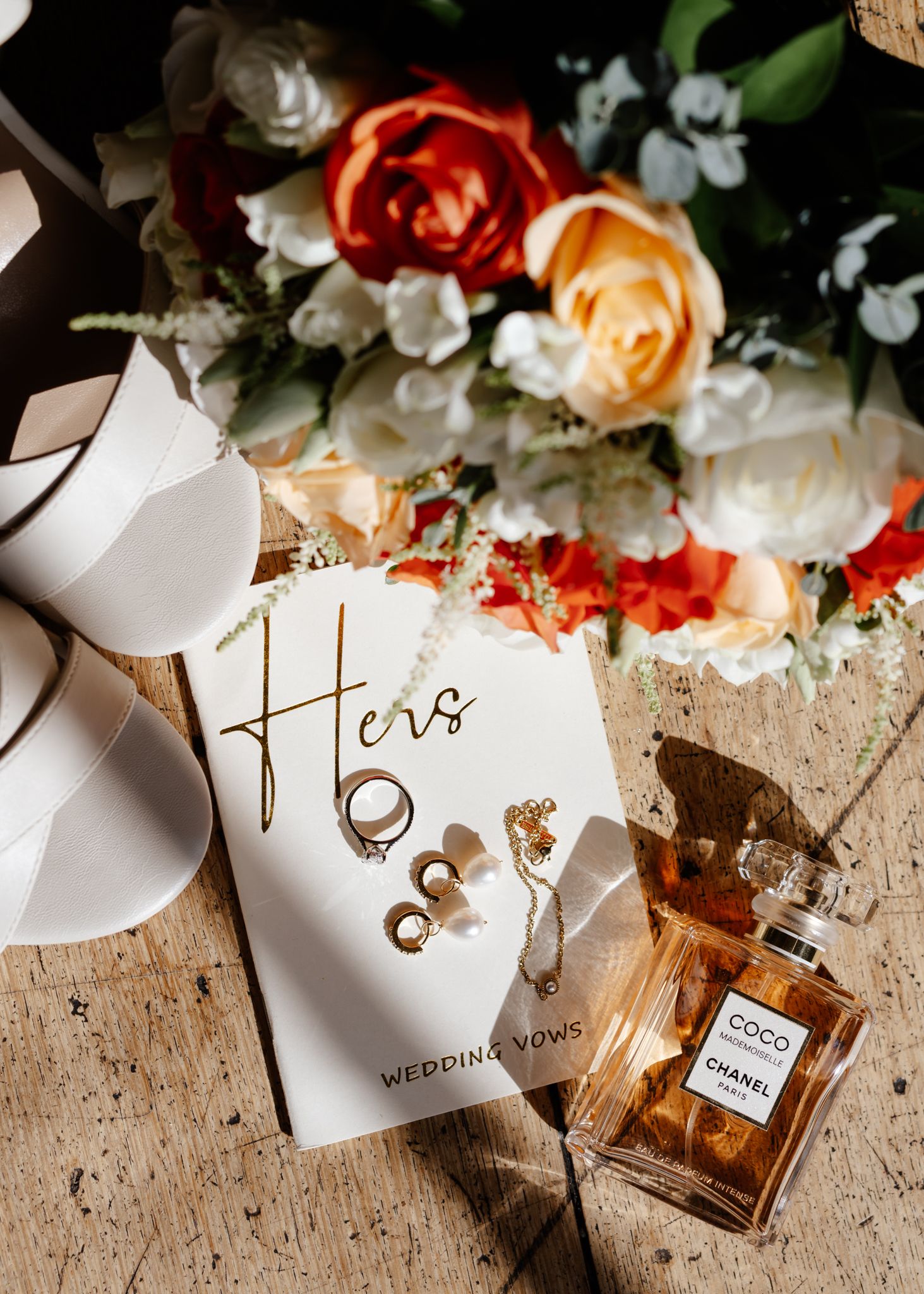 Wedding details flatlay with perfume bottle, name cards, and coral roses on rustic wooden surface.