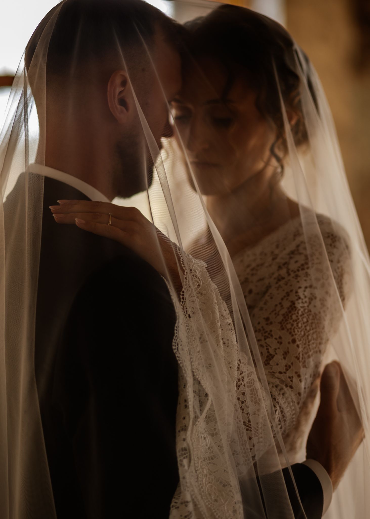 A romantic wedding couple shares an intimate moment under a delicate white veil in warm lighting.