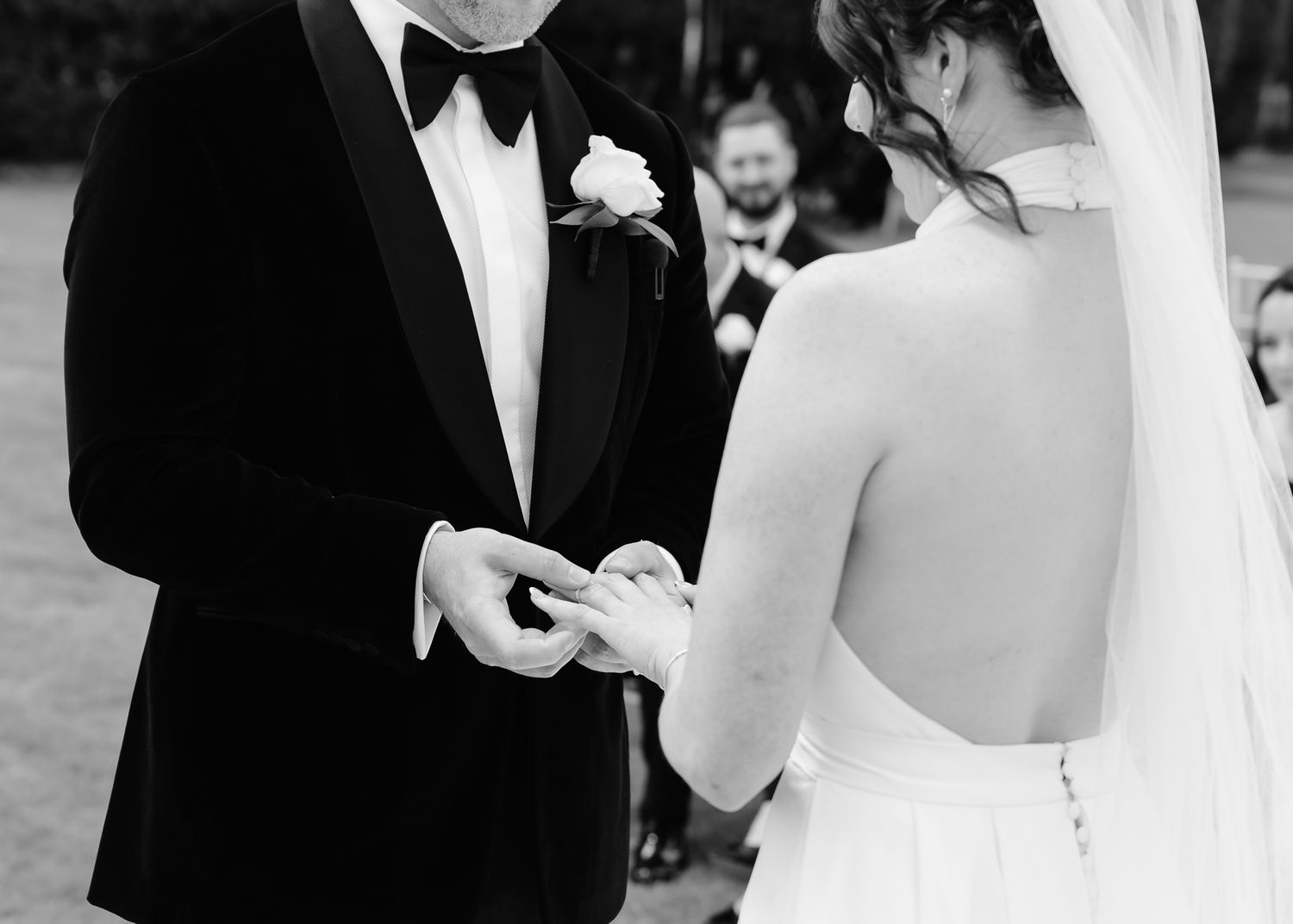Black and white photo of a wedding ceremony with elegant formal attire and ring exchange.