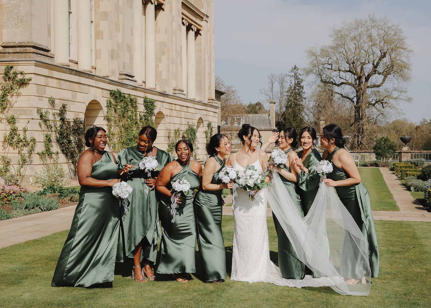 Wedding party in sage green bridesmaid dresses poses together on sunny lawn outside elegant stone building.