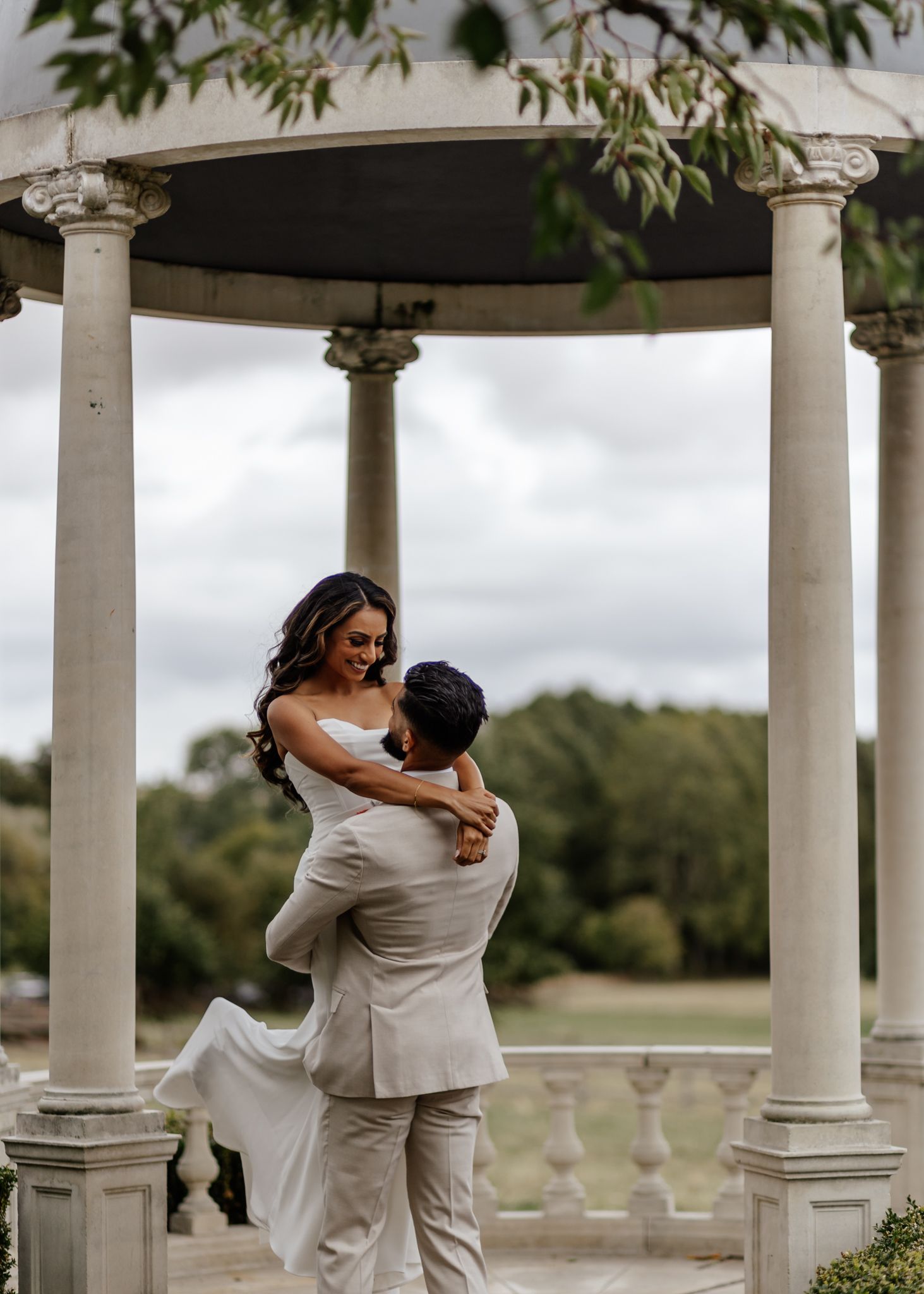 A couple in white outfits dances together in an elegant outdoor gazebo with white columns.