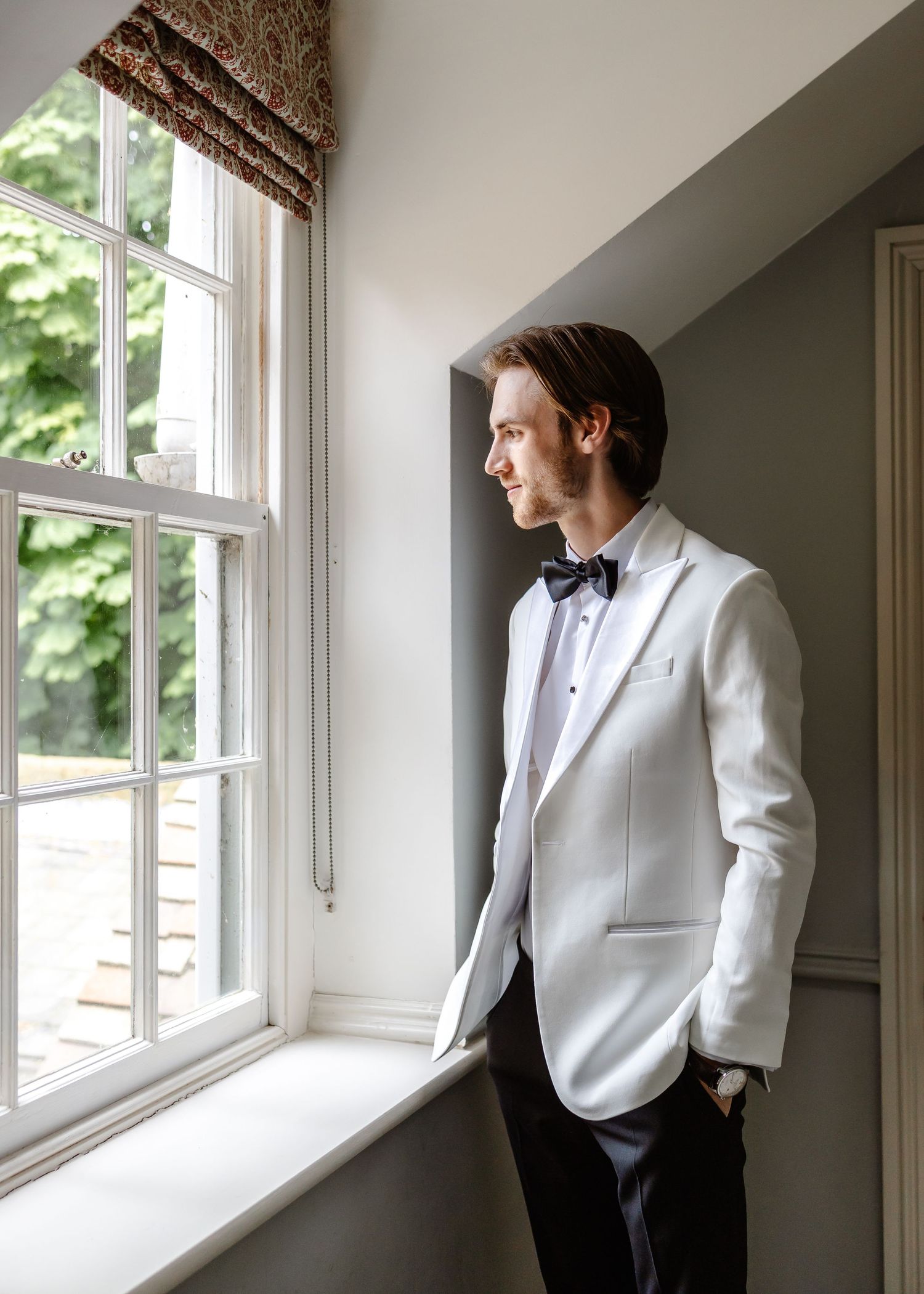 Groom in white tuxedo jacket and black bow tie gazes out window in elegant venue, natural light portrait.