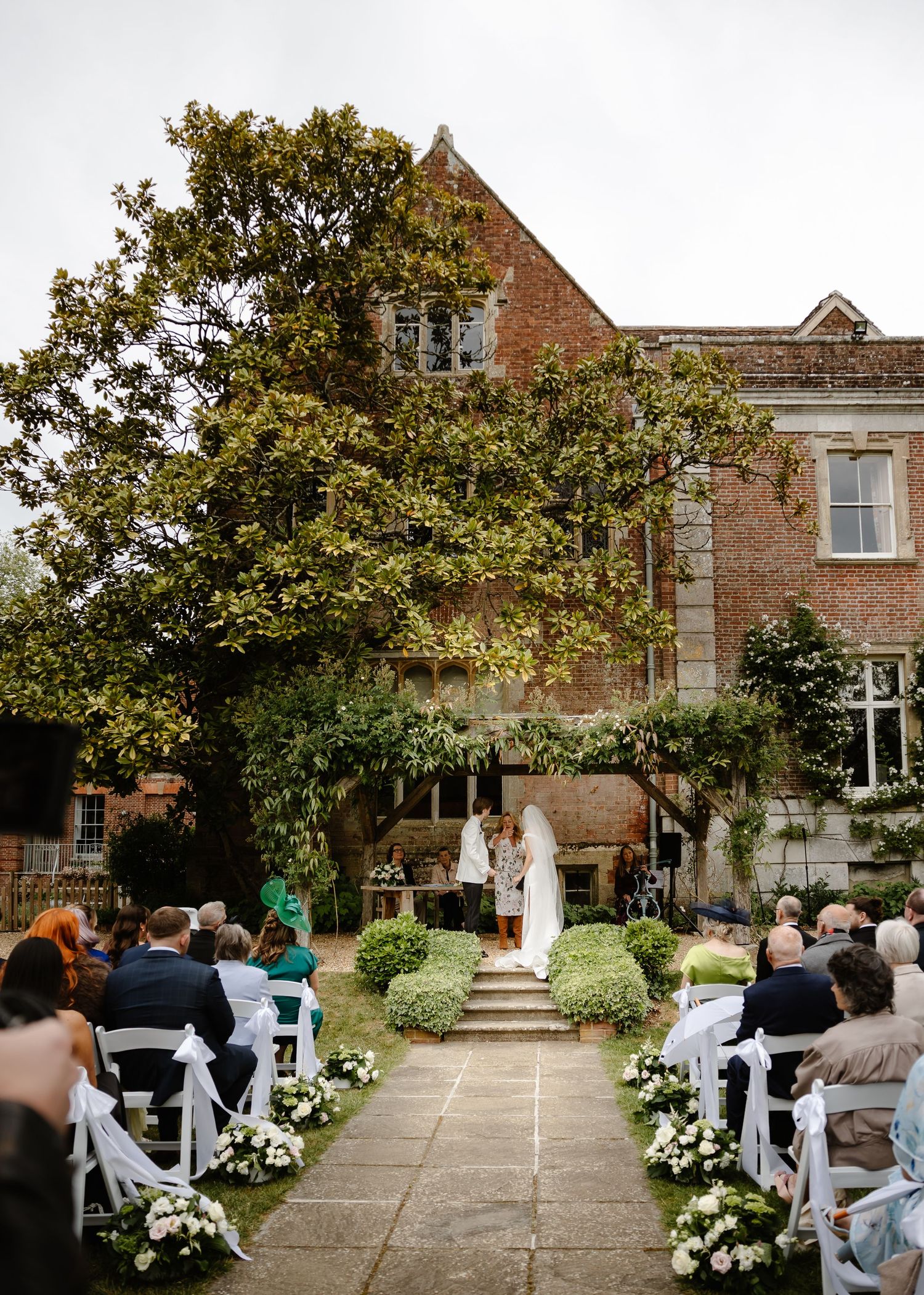 Outdoor wedding ceremony in front of a brick manor house with guests seated in white chairs along a floral aisle.