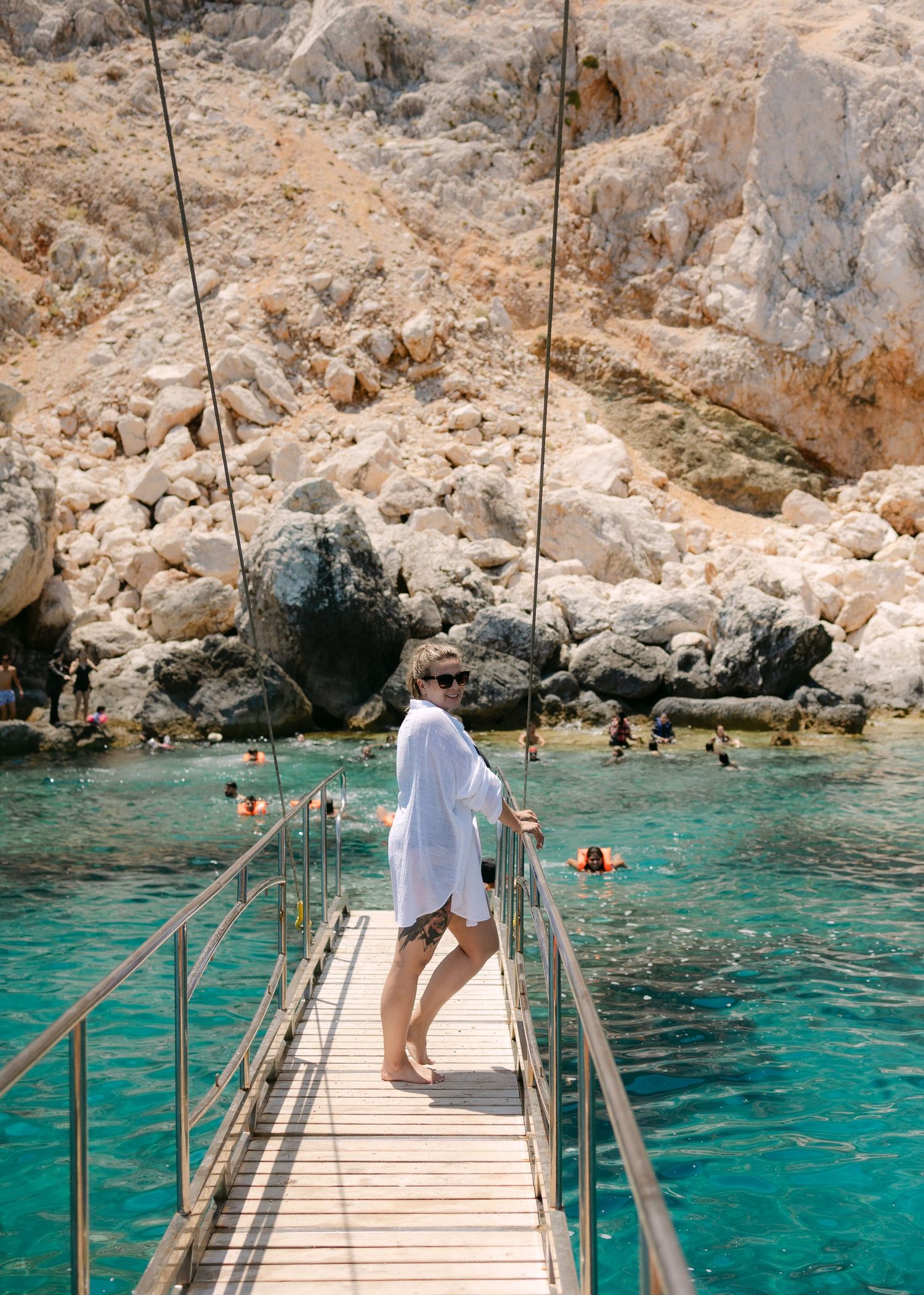 Woman in white shirt walks on a wooden bridge over turquoise water near rocky cliffs with swimmers in background.