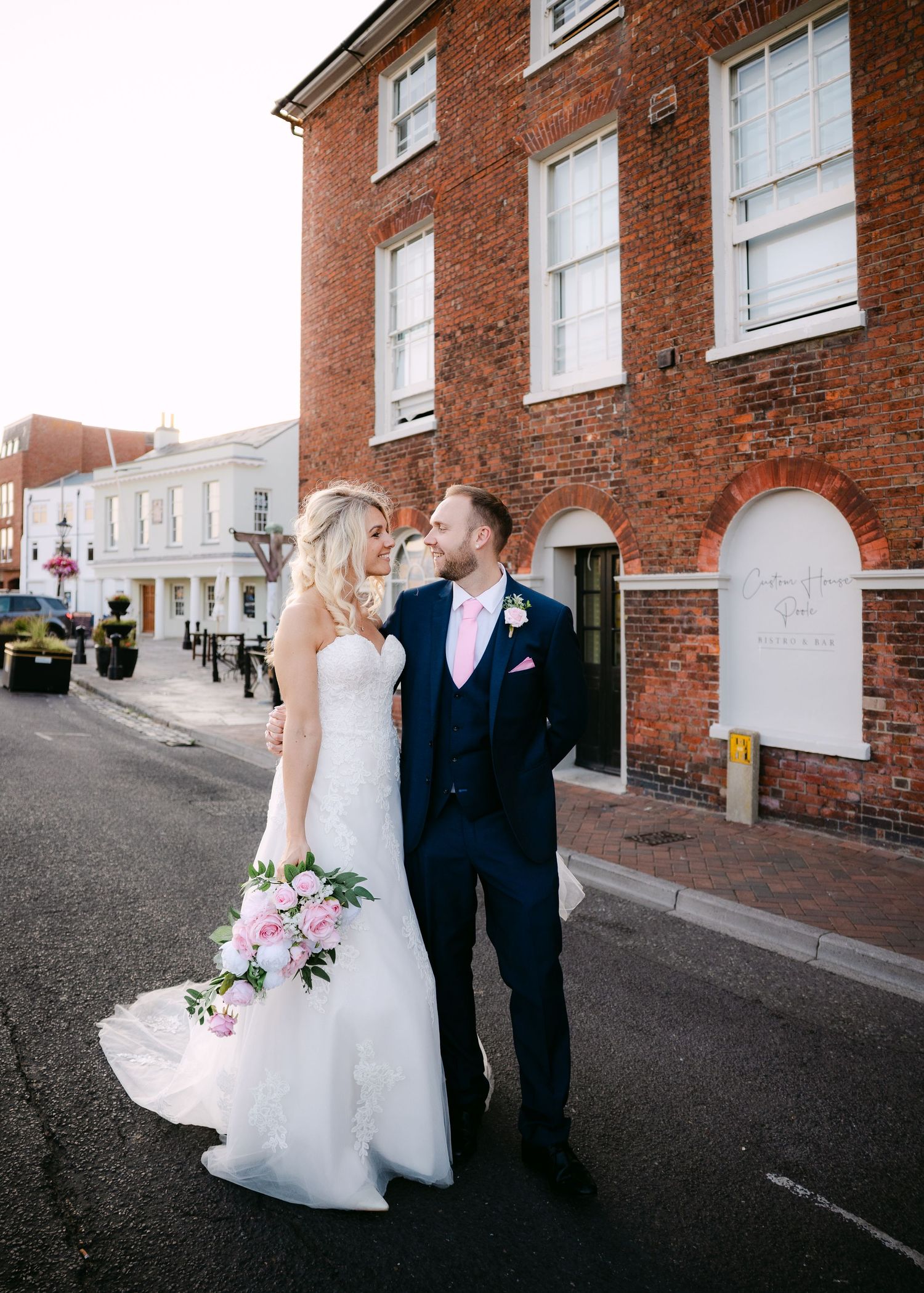 Bride and groom share a romantic moment on a street outside a red brick building, bride holding a pink bouquet.