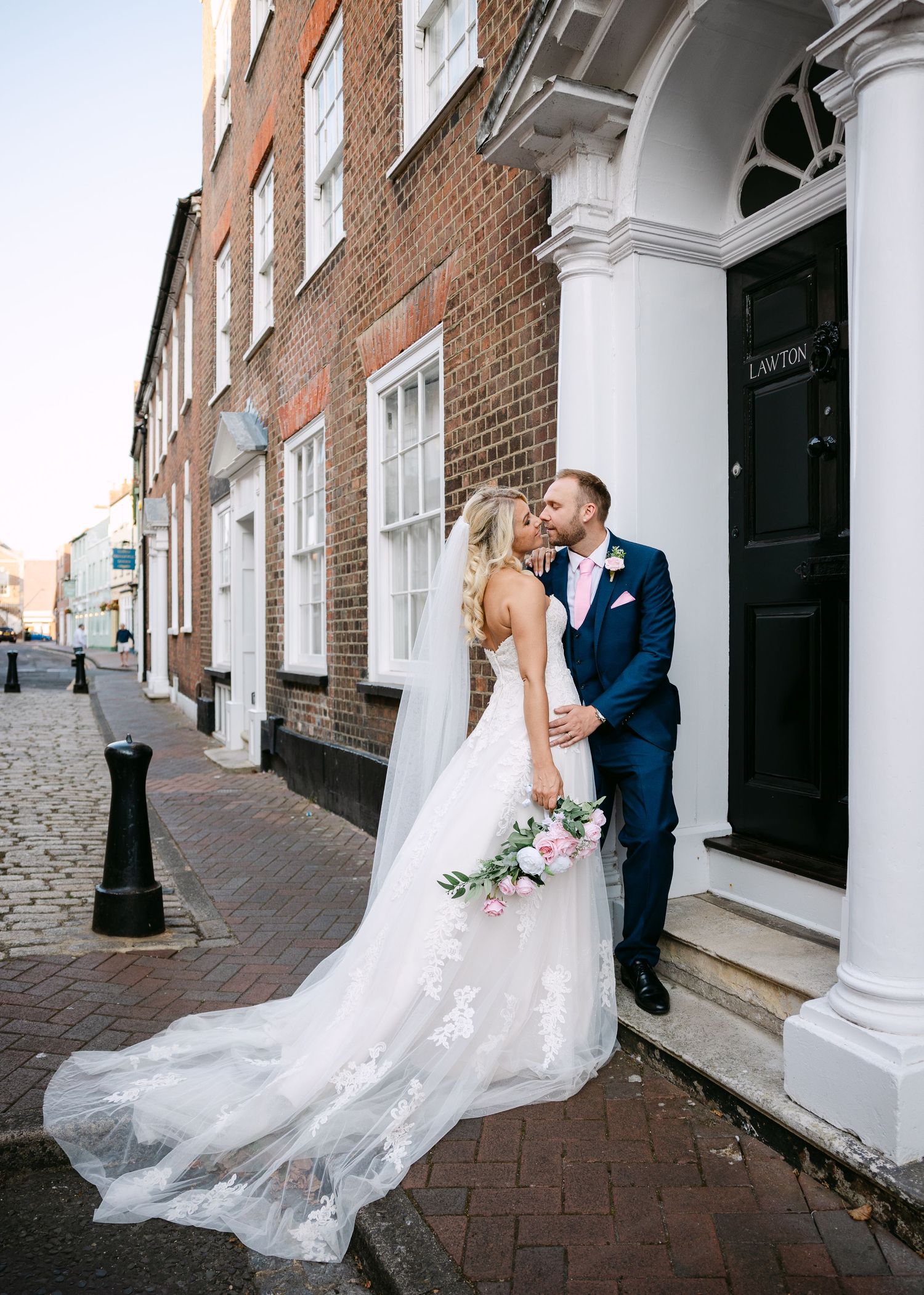 Bride and groom share a romantic kiss on a cobblestone street, bride in lace gown with long veil holding pink bouquet.