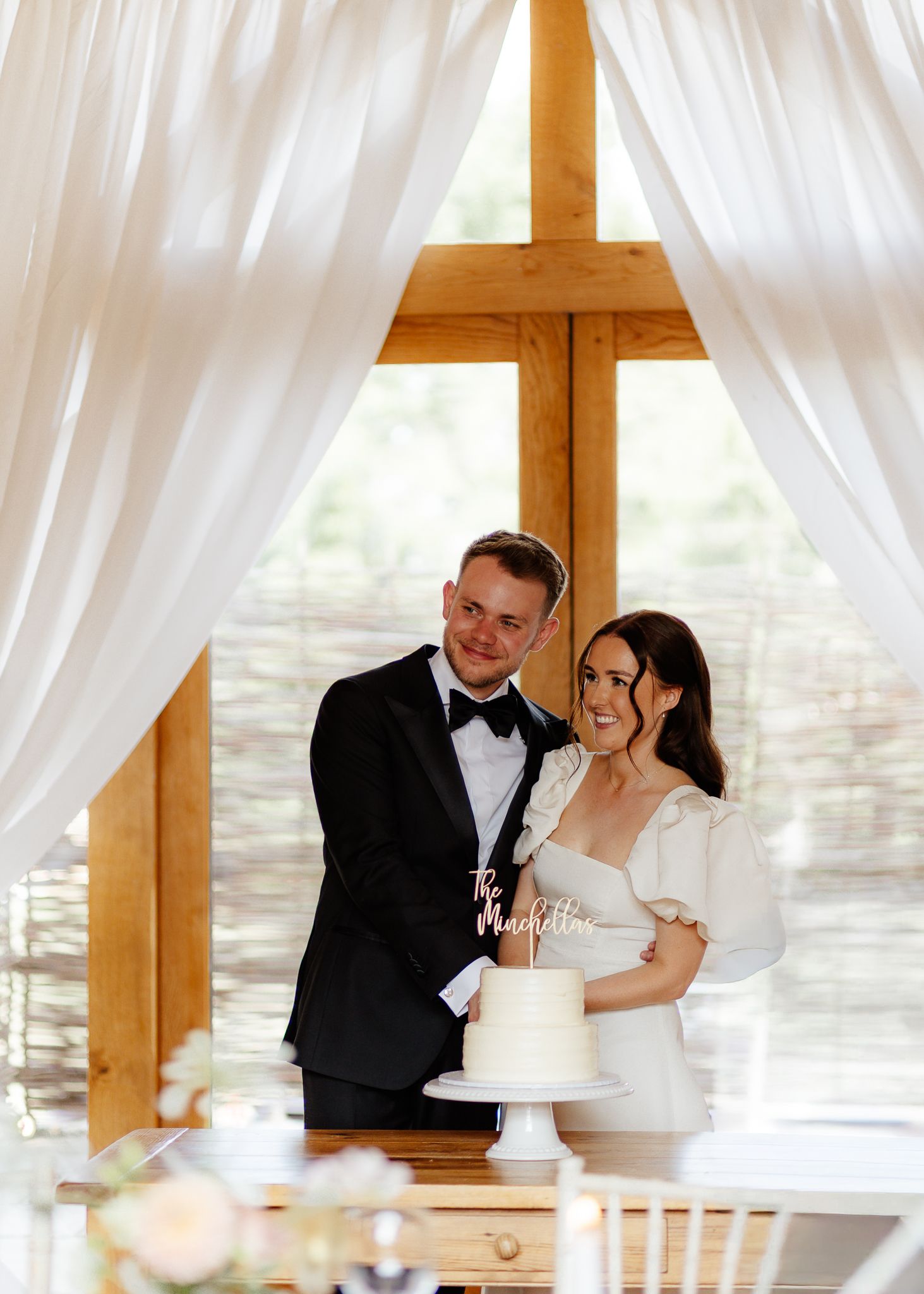 A couple shares an intimate moment during their wedding cake cutting ceremony under white drapes and wooden beams.