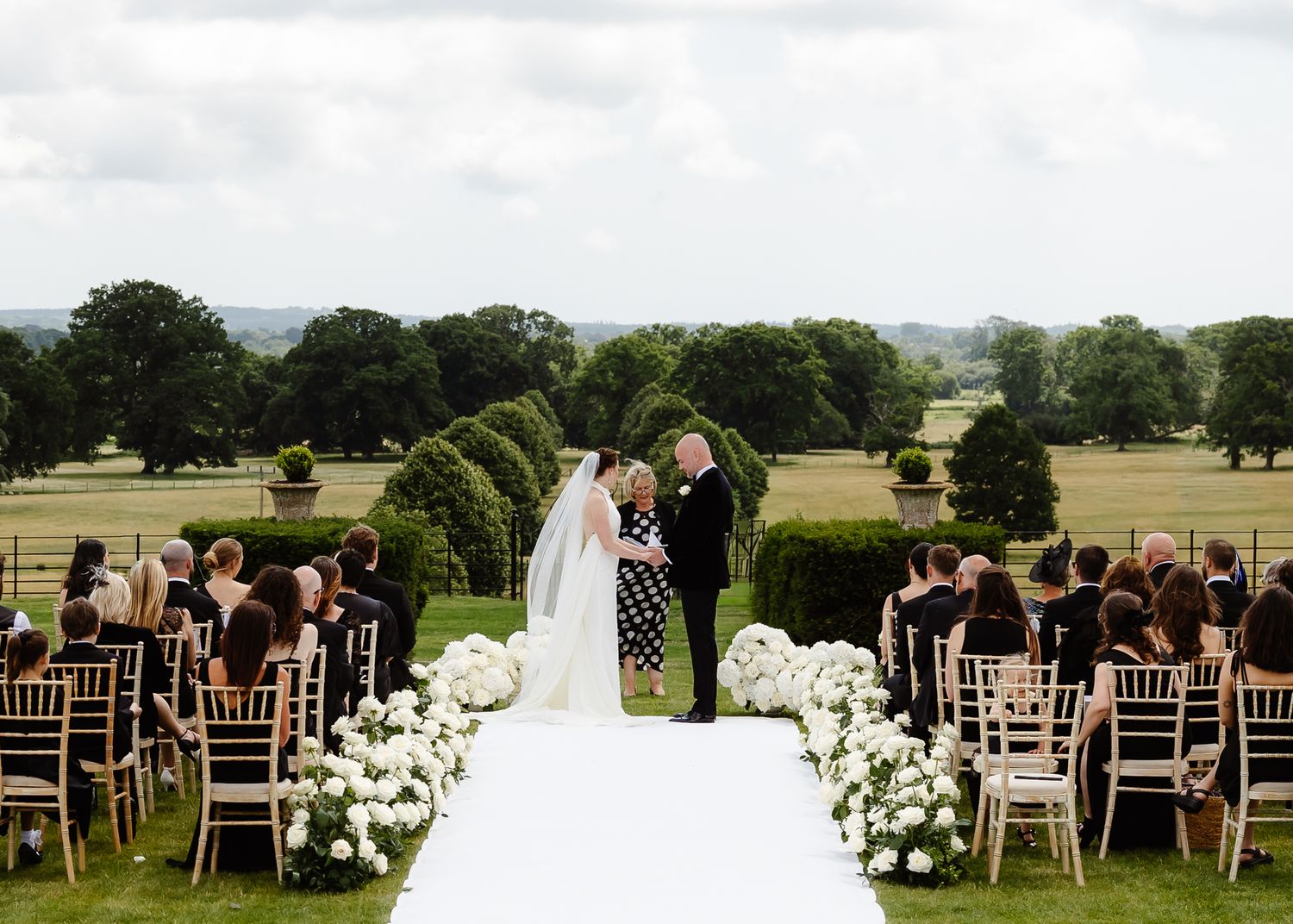 Outdoor wedding ceremony with white chairs and floral arrangements against a scenic countryside backdrop.