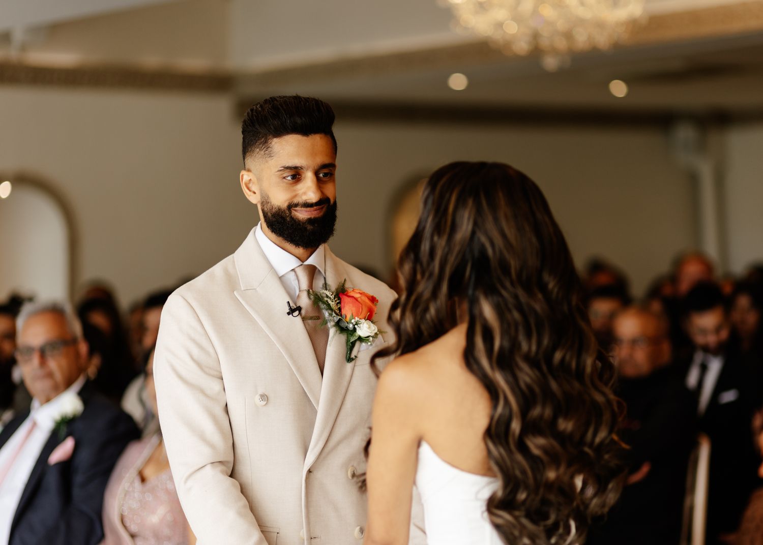 A couple exchanges vows during an indoor wedding ceremony with a warm-lit chandelier and guests in the background.