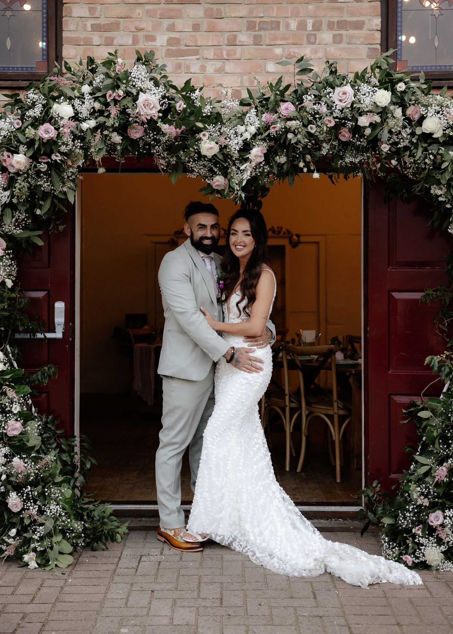 Romantic wedding couple poses in front of a rustic brick wall with white floral arrangements and string lights.
