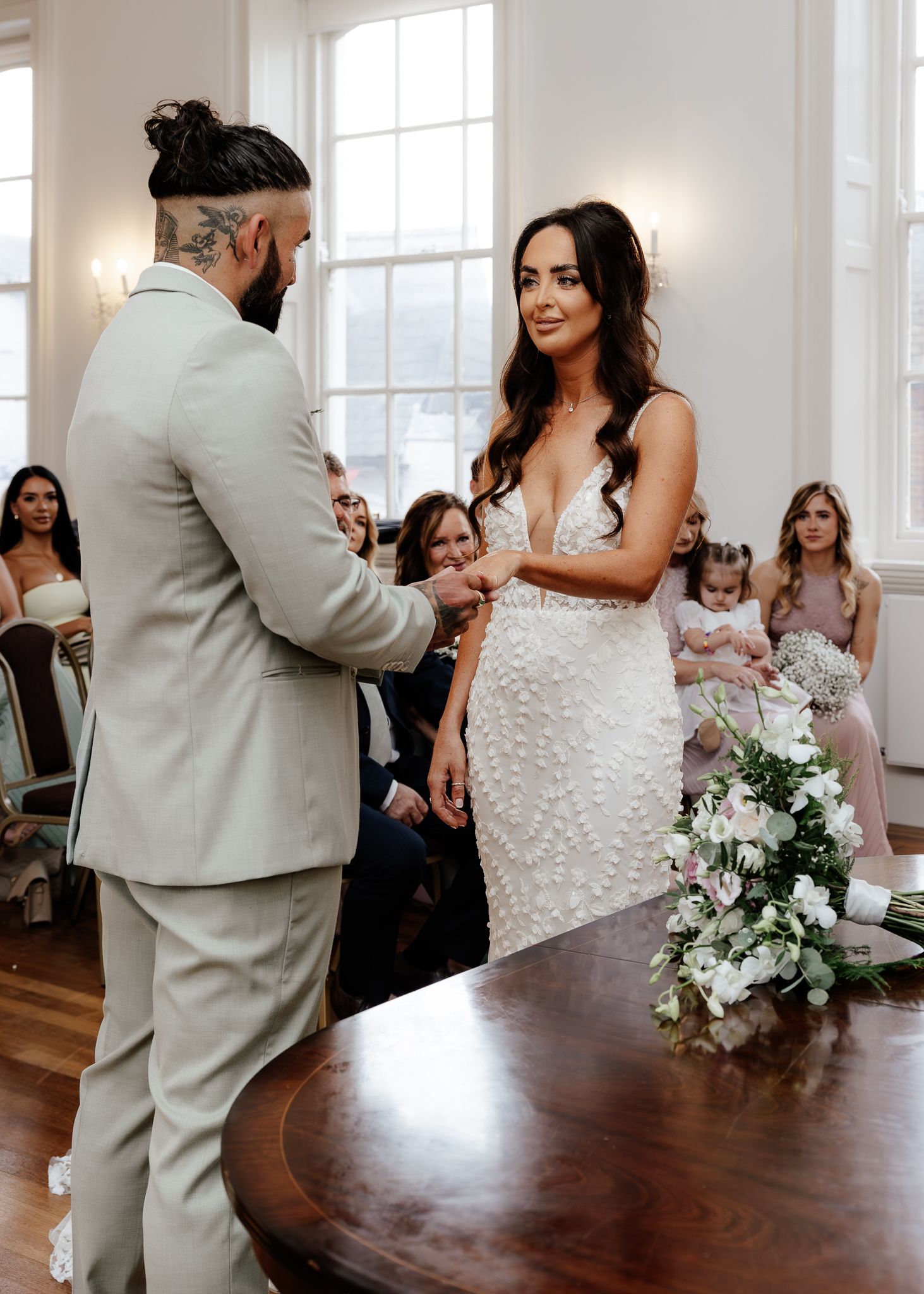 A couple shares their first dance in a bright white room with large windows and a dark wooden table in the foreground.